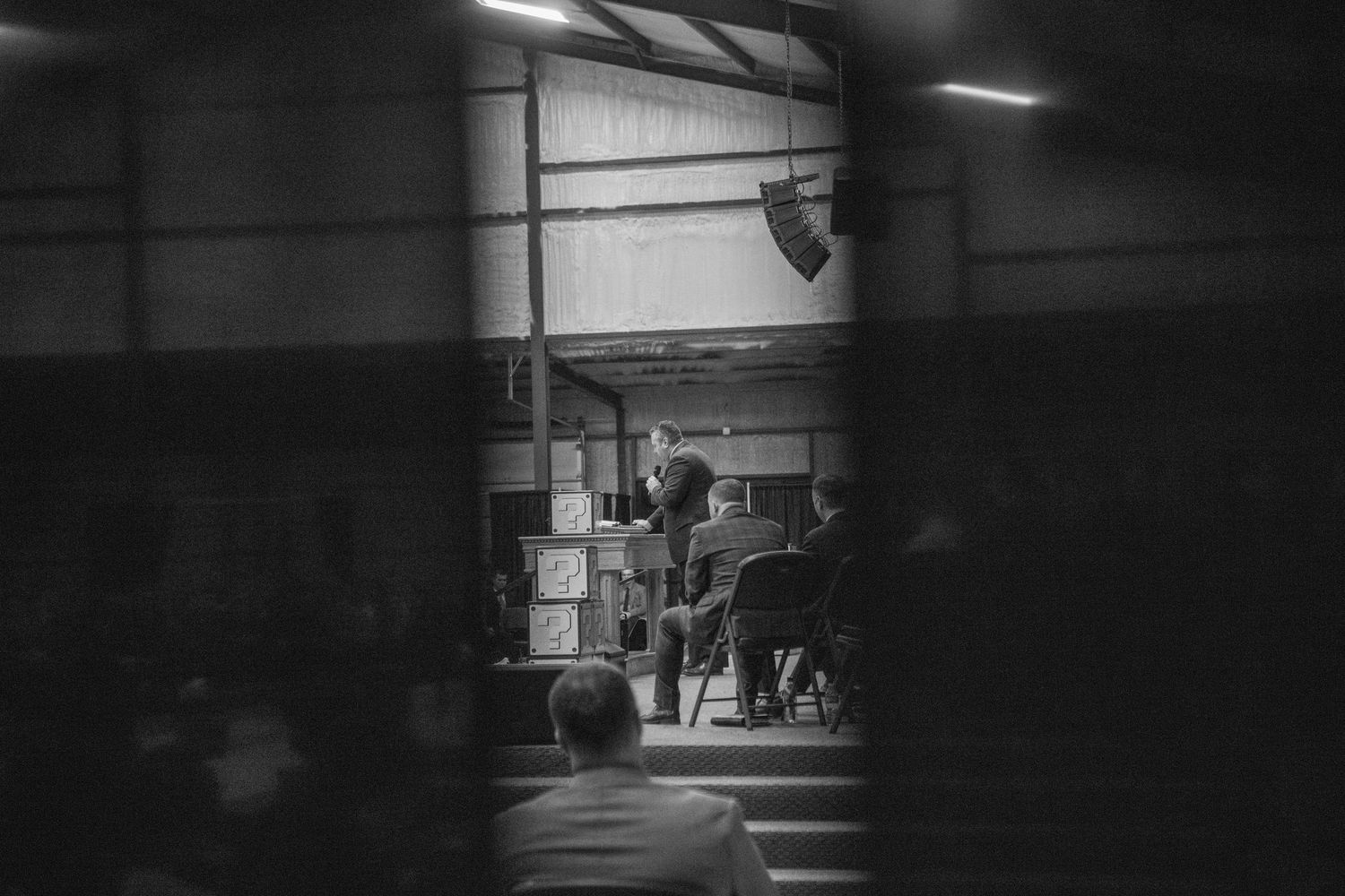 Black and white view through a window of a preacher delivering a sermon to a seated congregation.