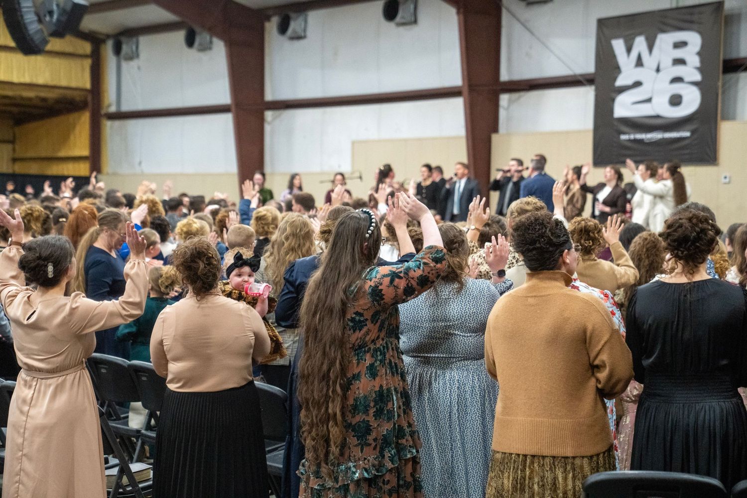 Large congregation of women raising hands in worship at WR26 church conference event.