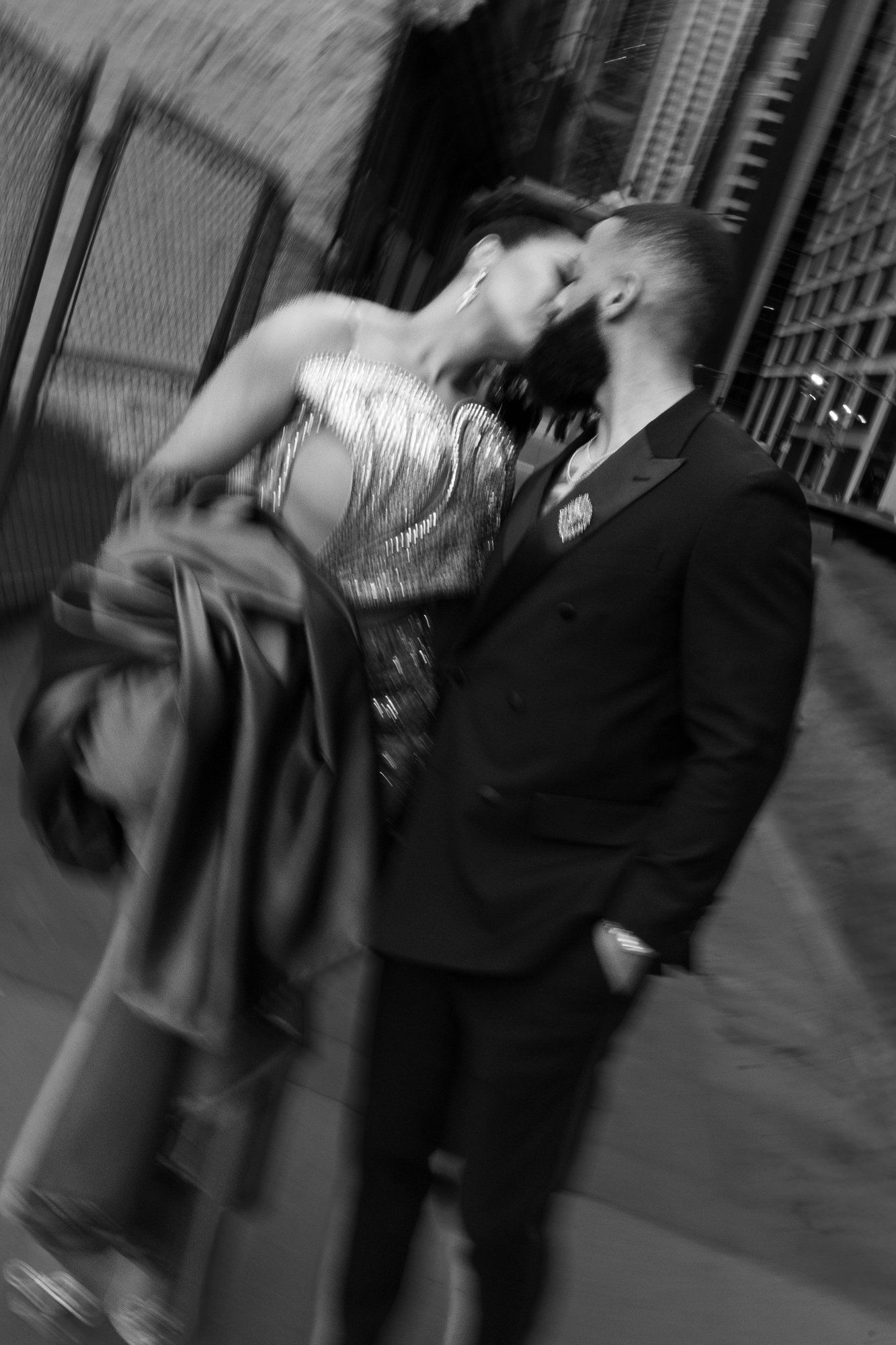 Black and white photo of a couple kissing in an alley, woman in sequin dress, man in suit, with artistic motion blur.