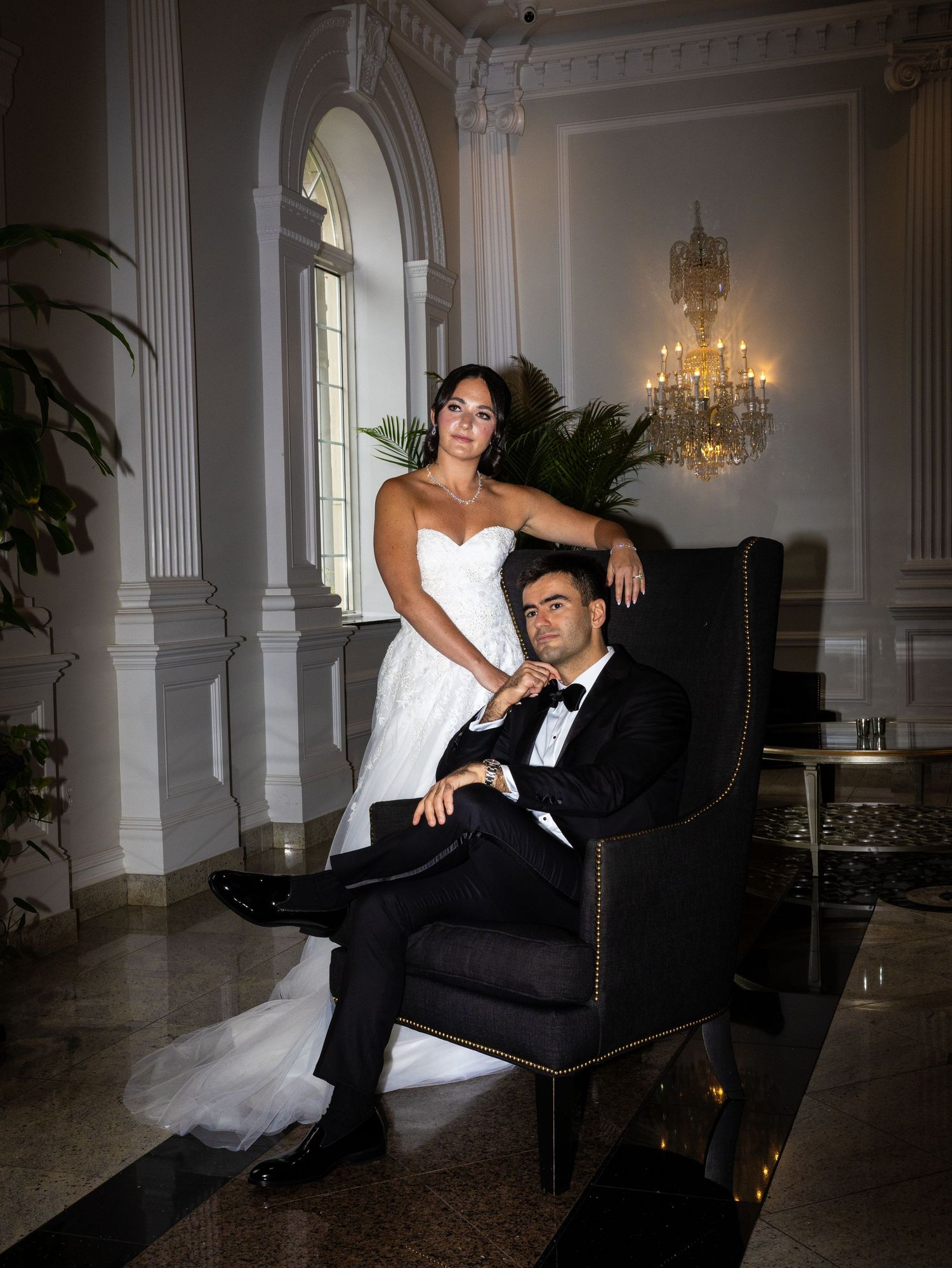 Elegant bride and groom posing in grand ballroom with chandelier, bride standing behind seated groom in New Jersey.