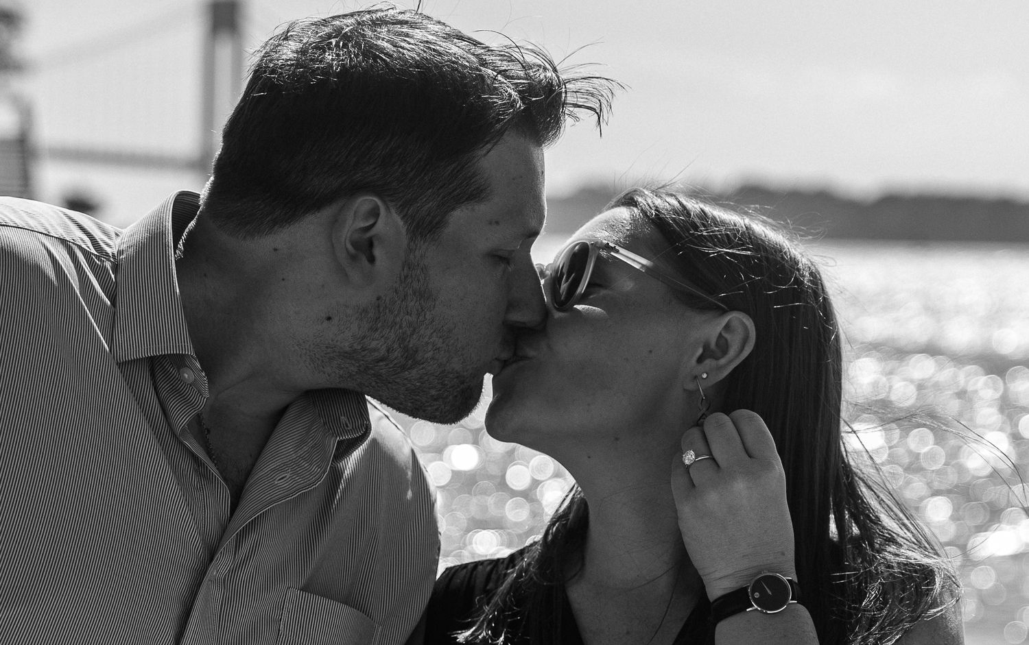 Black and white photo of a couple sharing a romantic kiss outdoors near a bridge and waterfront.