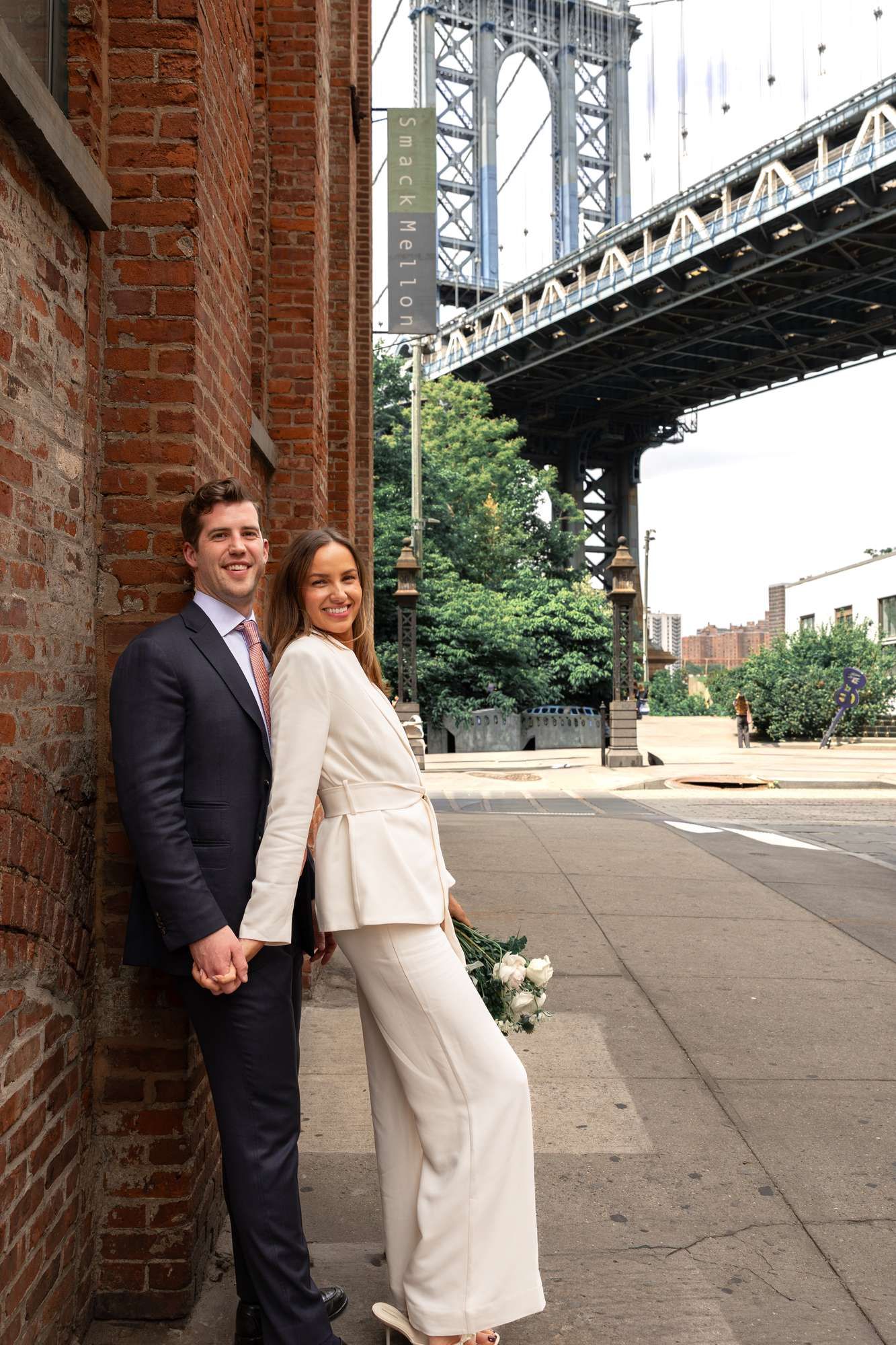 Newlyweds holding hands by brick wall in DUMBO Brooklyn with Manhattan Bridge visible behind them.
