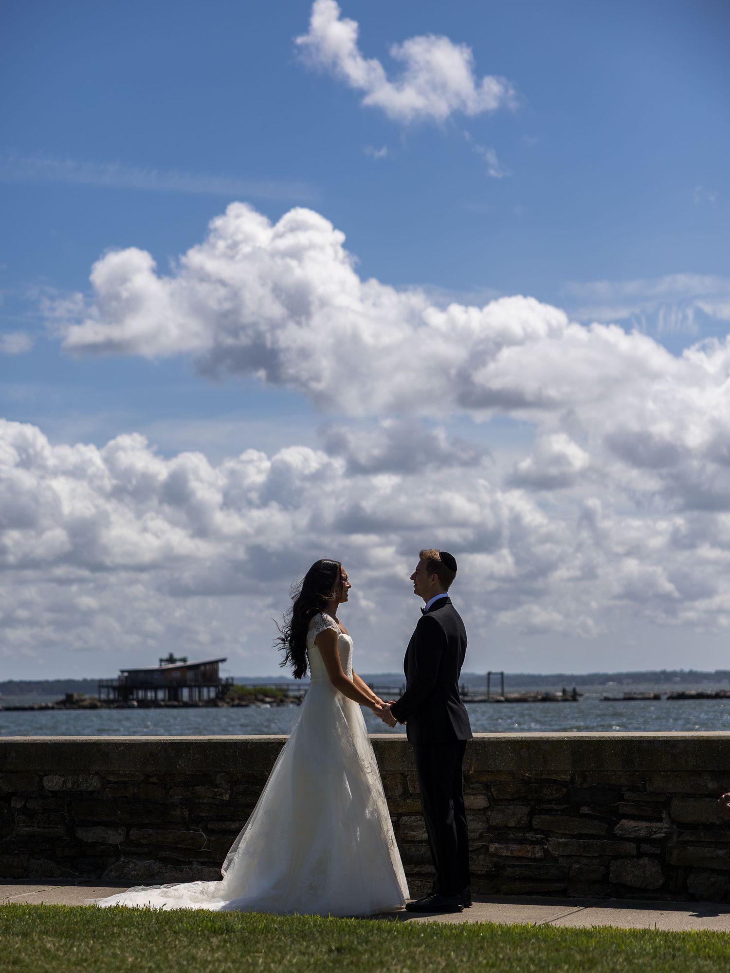 Bride and groom hold hands facing each other by waterfront under dramatic cloudy sky in Glen Island, Bronx, New York City