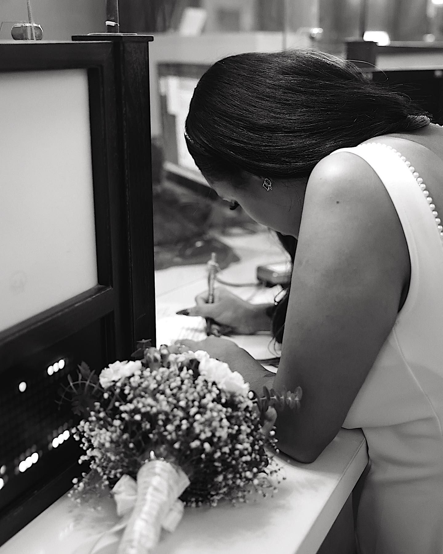 Black and white photo of a bride in a white dress signing documents with a bridal bouquet on the counter beside her.