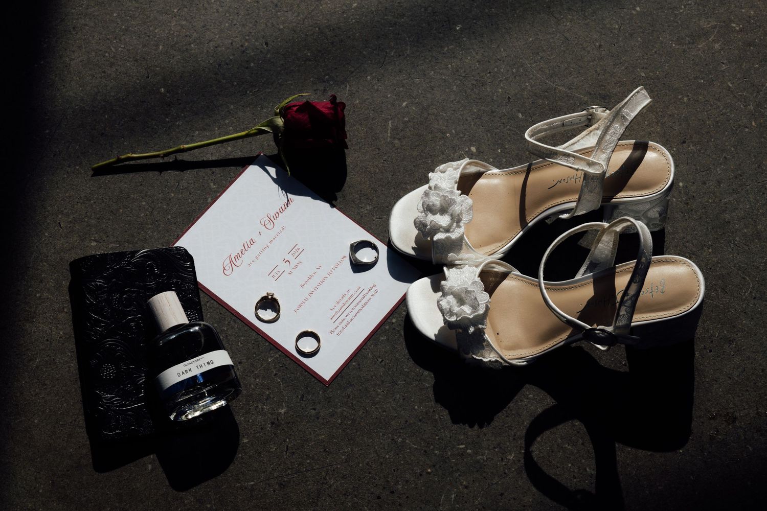 Bridal flat lay with white floral sandals, wedding invitation, two rings, a red rose and perfume bottle on dark pavement.