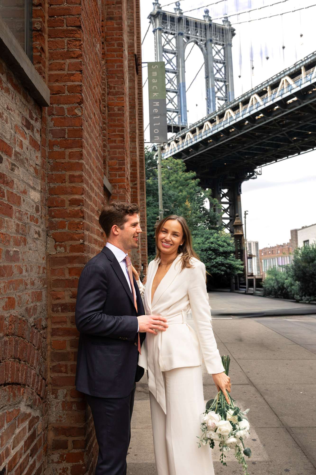 Bride in white pantsuit holding bouquet stands with groom in navy suit near Manhattan Bridge in DUMBO, Brooklyn.