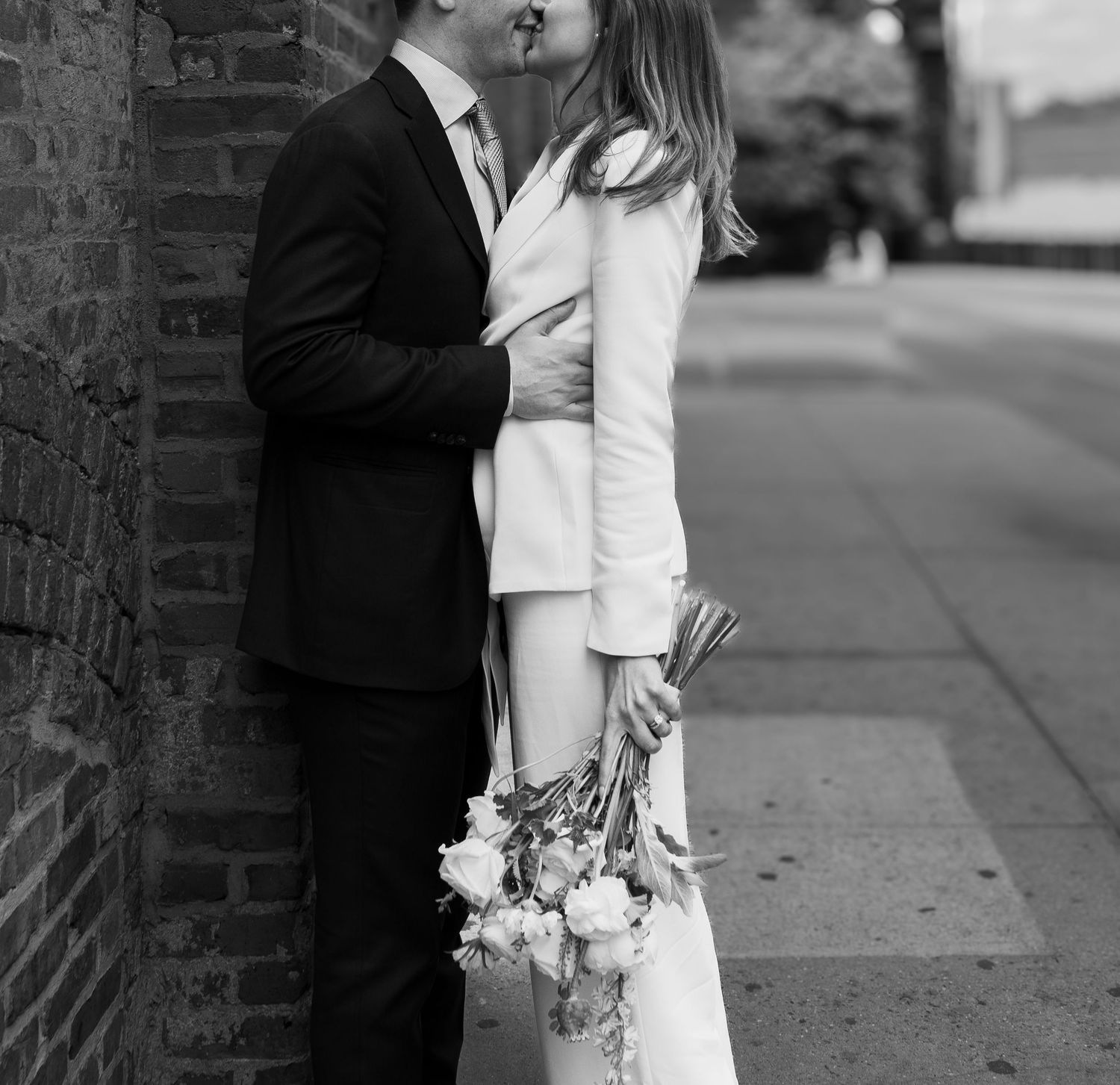 Black and white photo of a couple kissing on a city sidewalk, bride holding a bouquet in a white suit.
