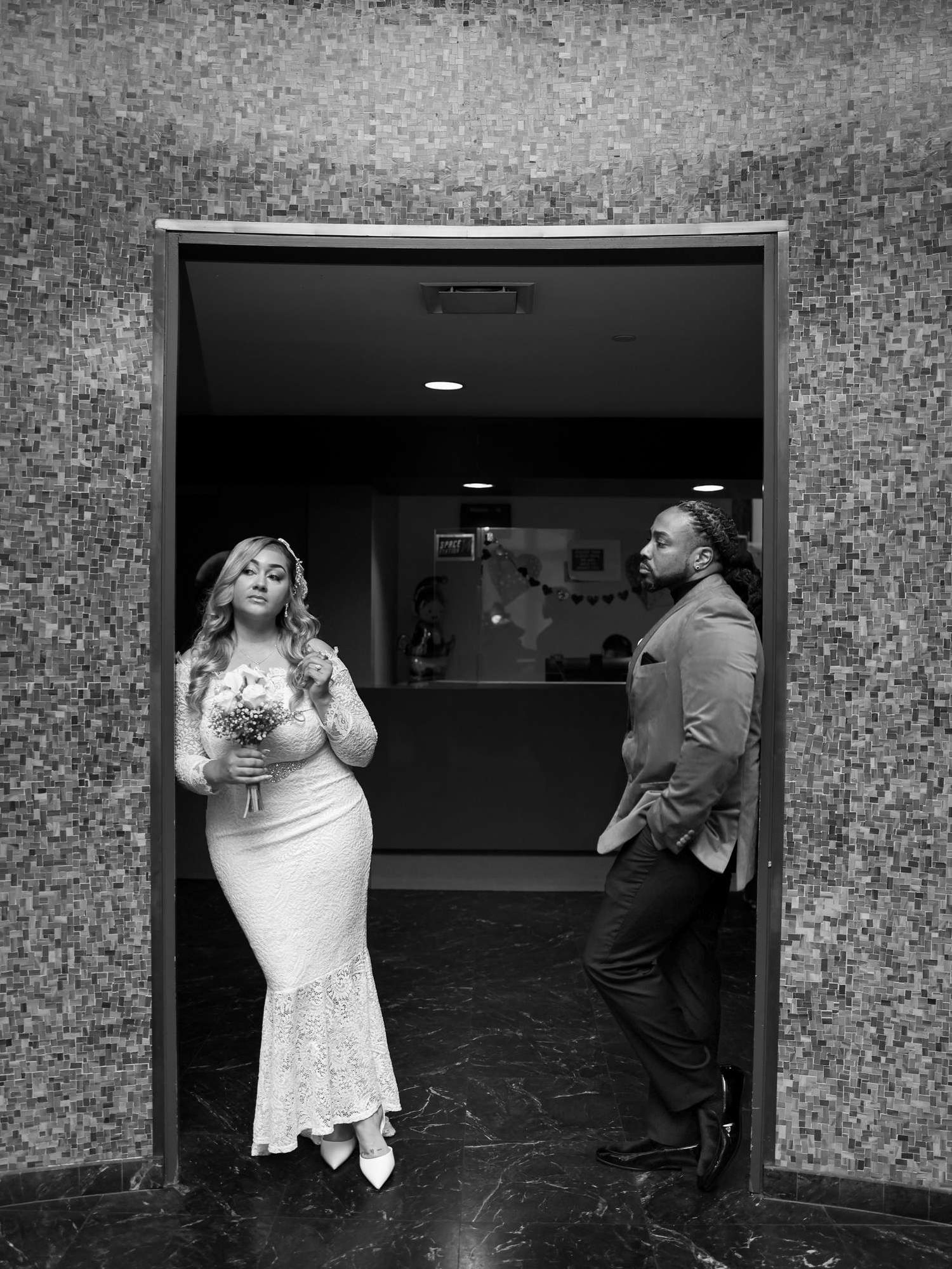Black and white photo of a bride in white dress holding bouquet and groom in suit standing in a doorway.