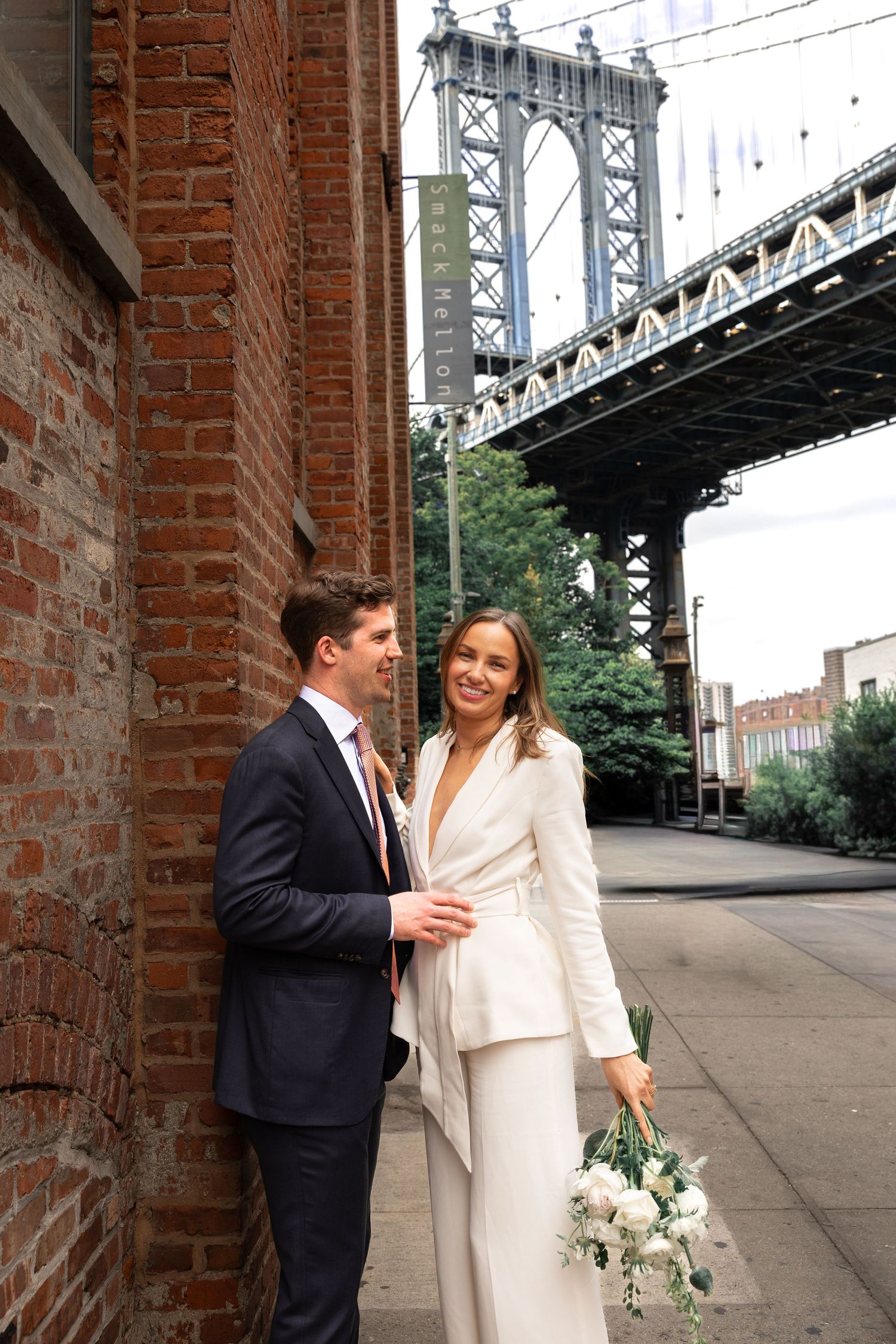 Bride in white pantsuit holding bouquet with groom in navy suit near Manhattan Bridge in DUMBO, Brooklyn.