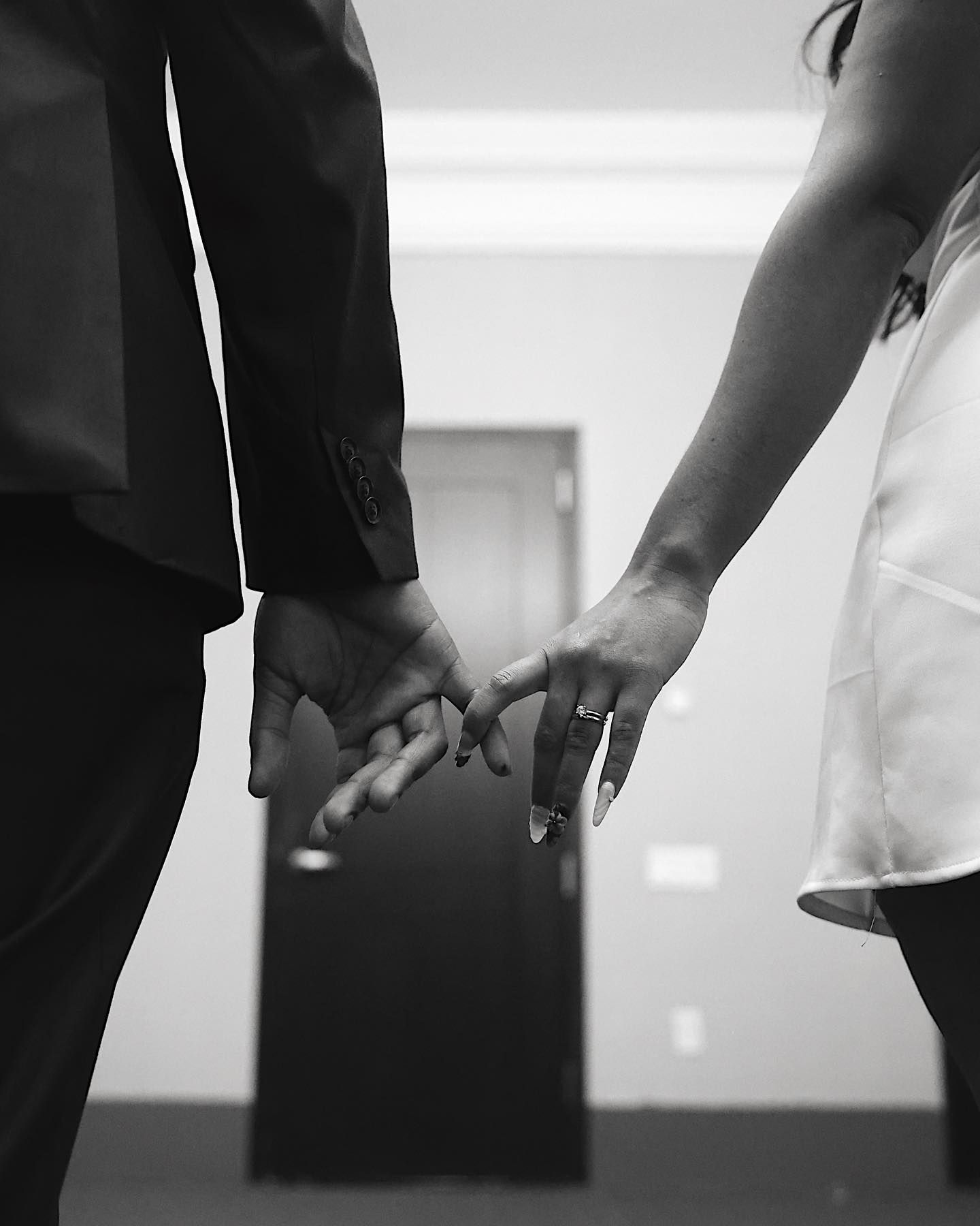 Black and white photo of a couple holding hands, woman wearing an engagement ring, dressed formally.