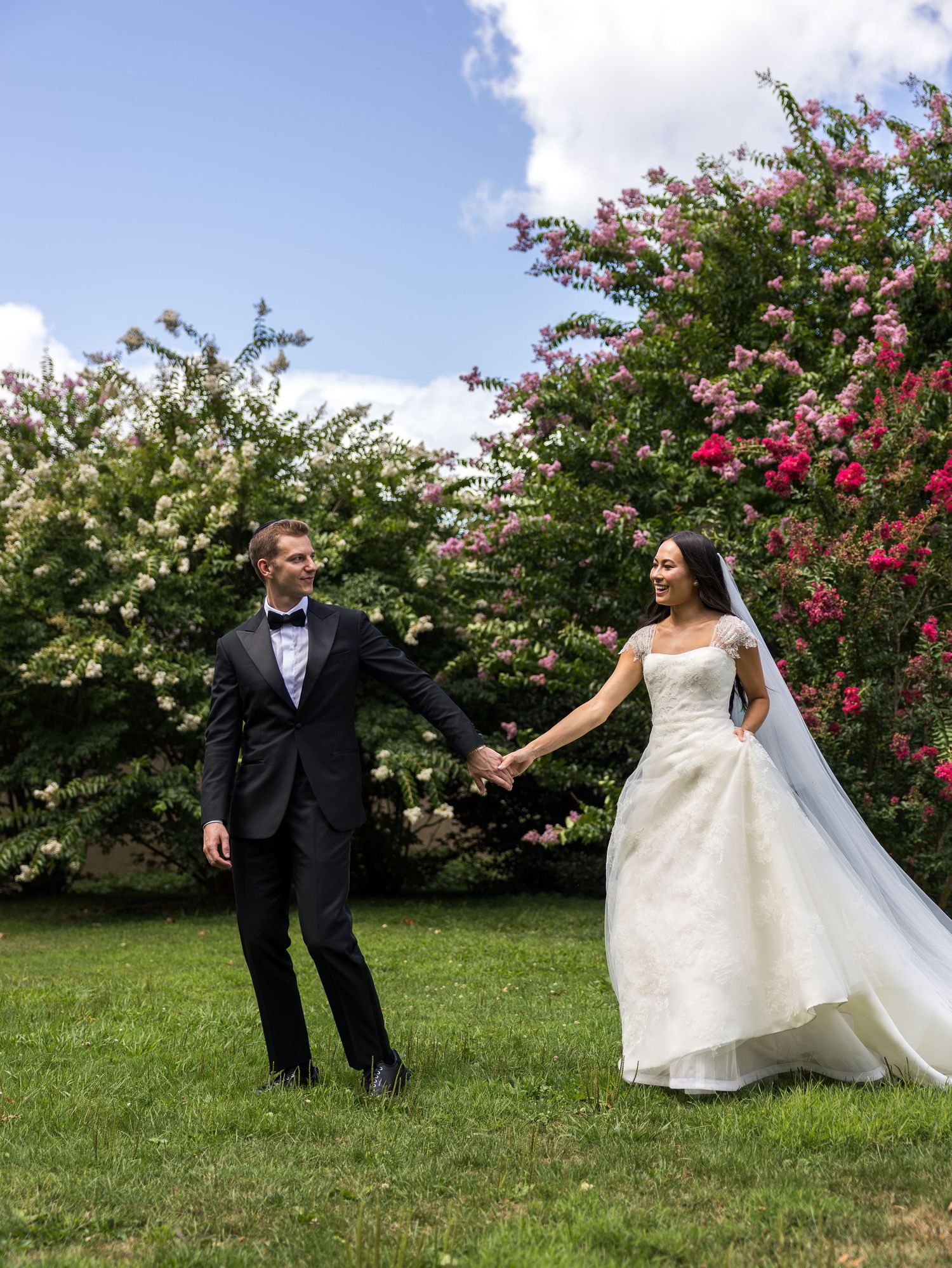 Bride and groom hold hands in lush garden with blooming pink and white trees, in Glen Island, New York City in the Bronx.