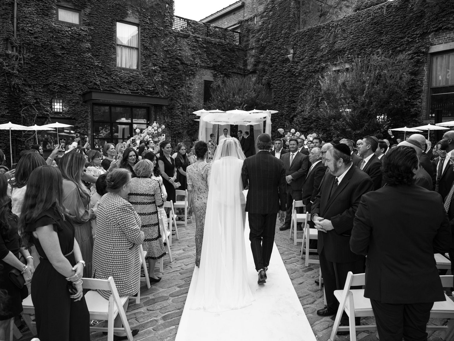 Black and white photo of a bride walking down the aisle with guests cheering at an outdoor wedding ceremony in The Foundry, Long Island City
