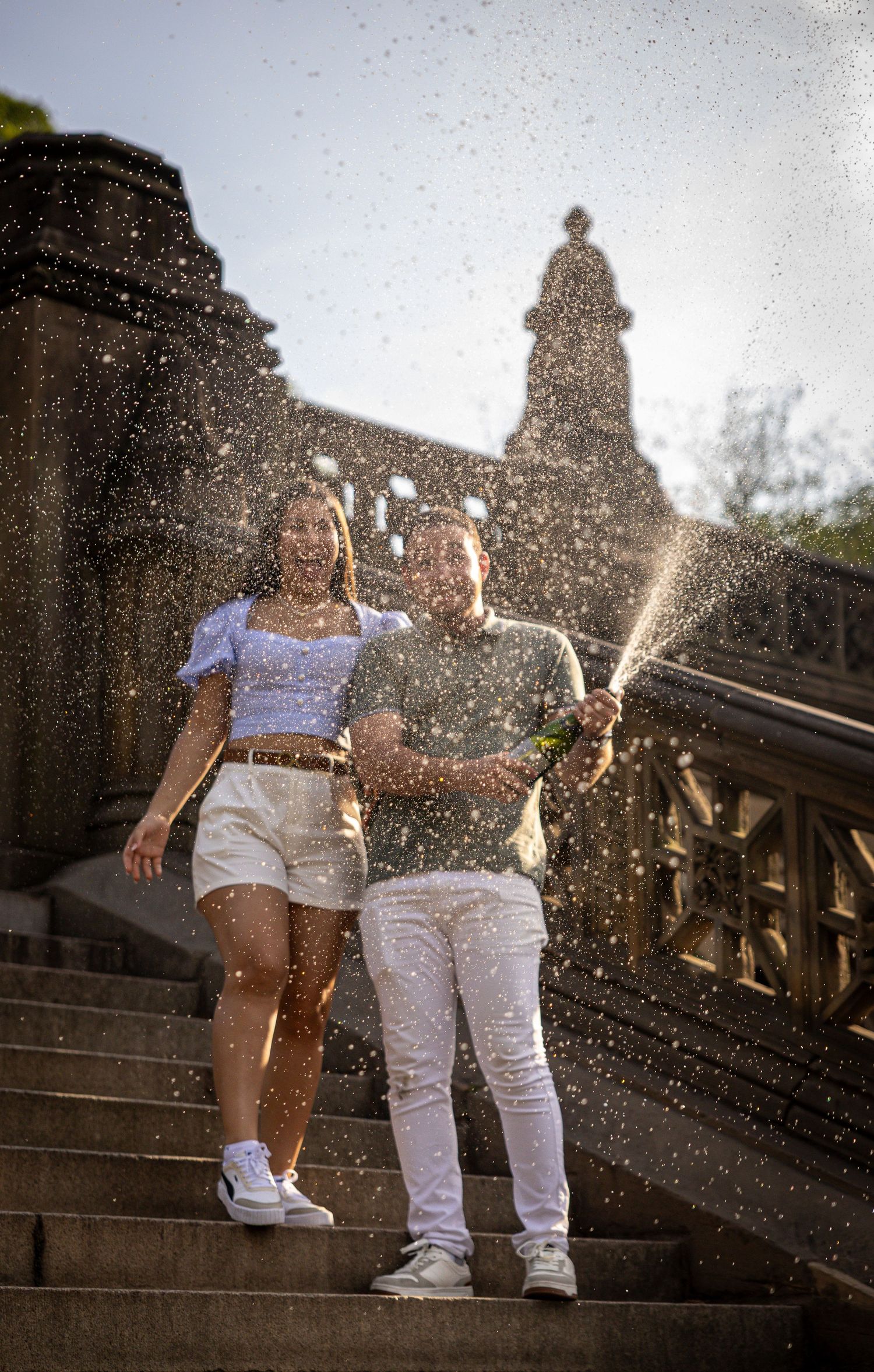 Couple celebrating on stone steps, spraying champagne in sunlight with a historic building backdrop.