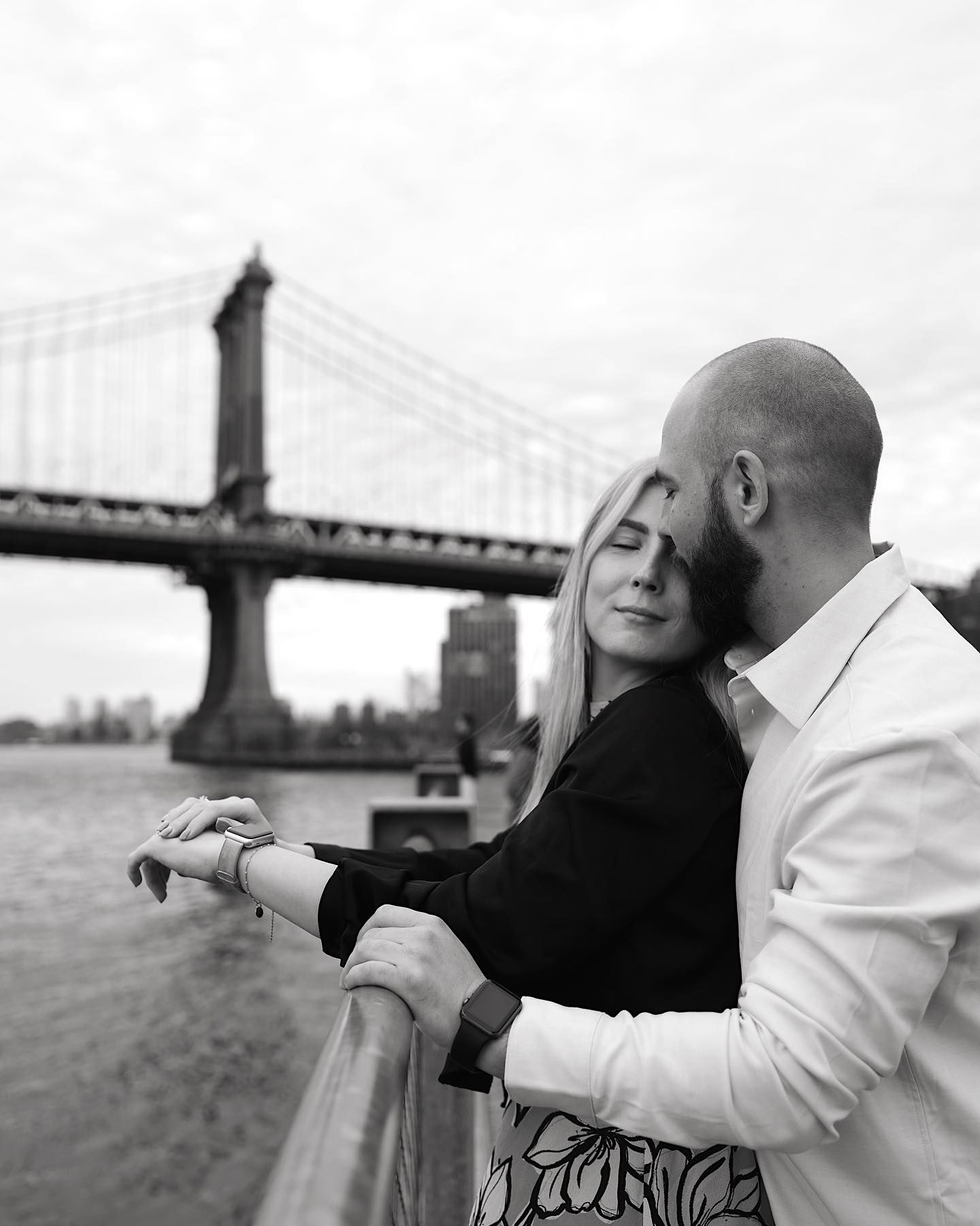 Black and white photo of a couple embracing near a railing with the Manhattan Bridge in the background.