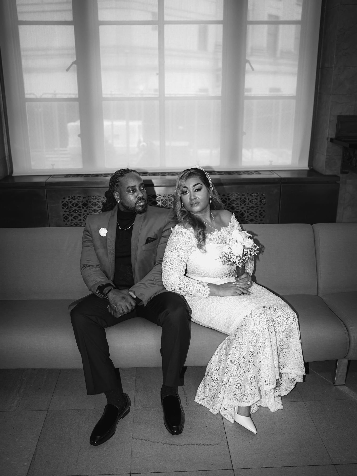 Black and white wedding photo of a couple sitting on a bench, bride in lace gown holding bouquet, groom in suit.