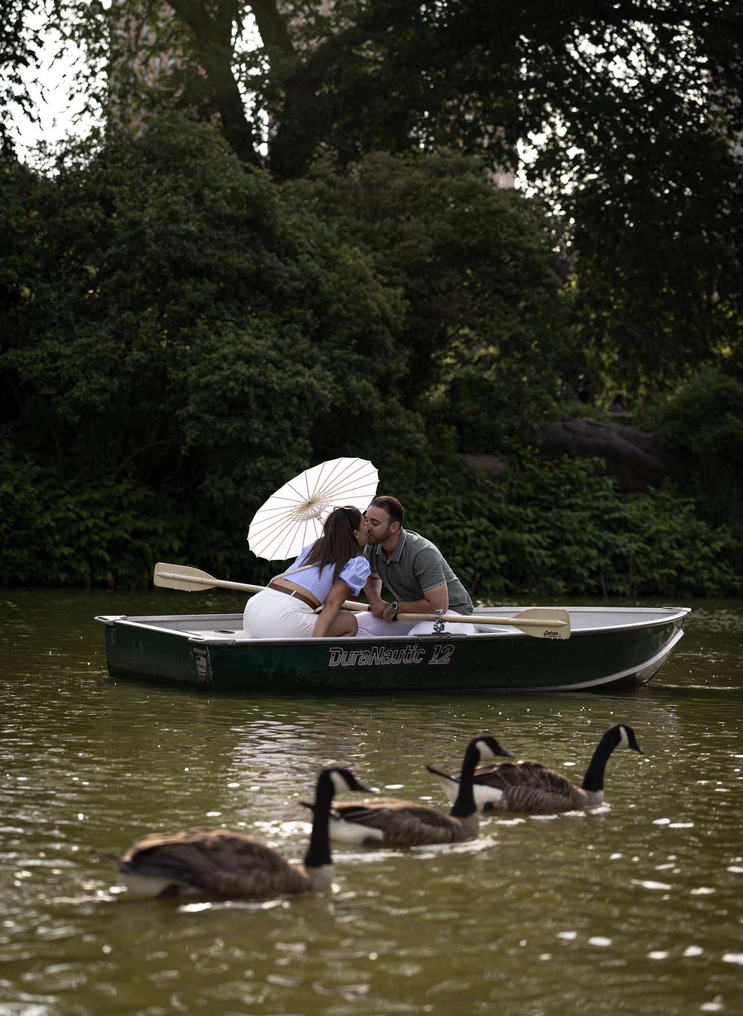 Couple sharing a romantic kiss in a rowboat on a lake, holding a parasol, with Canada geese swimming nearby.