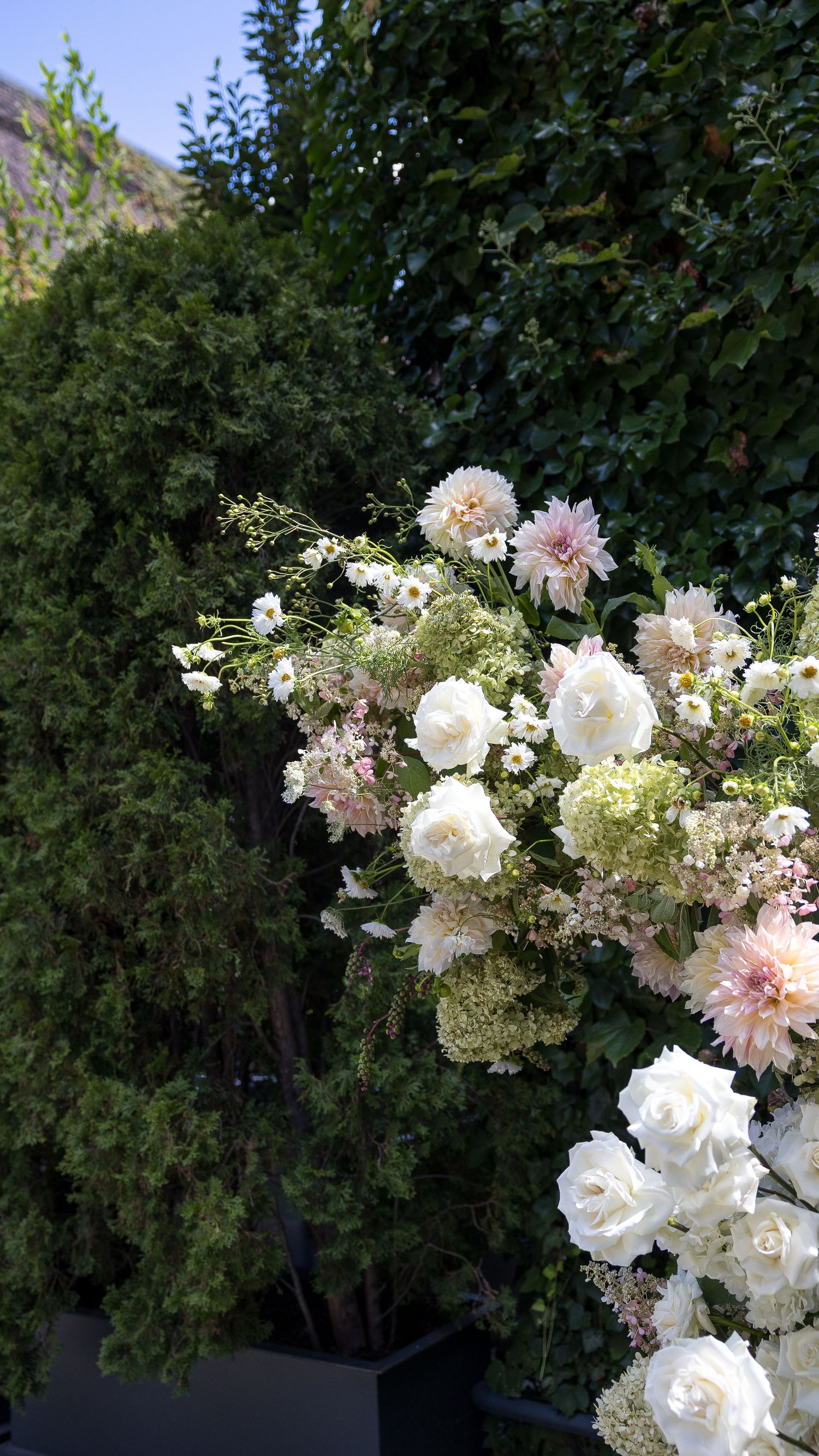 Lush white and blush floral arrangement with roses, dahlias, and wildflowers against green garden trees. In the Foundry, Long Island City