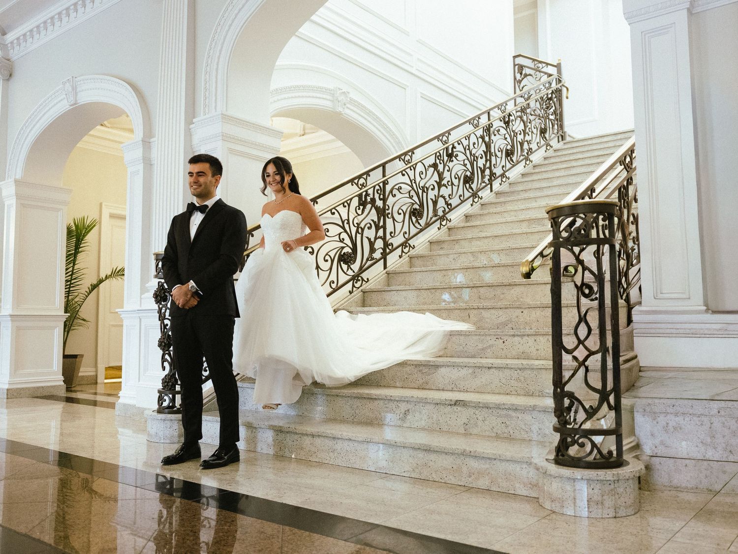 Elegant bride and groom posing on grand marble staircase with ornate railing in luxurious white wedding venue in The Rockeigh New jersey