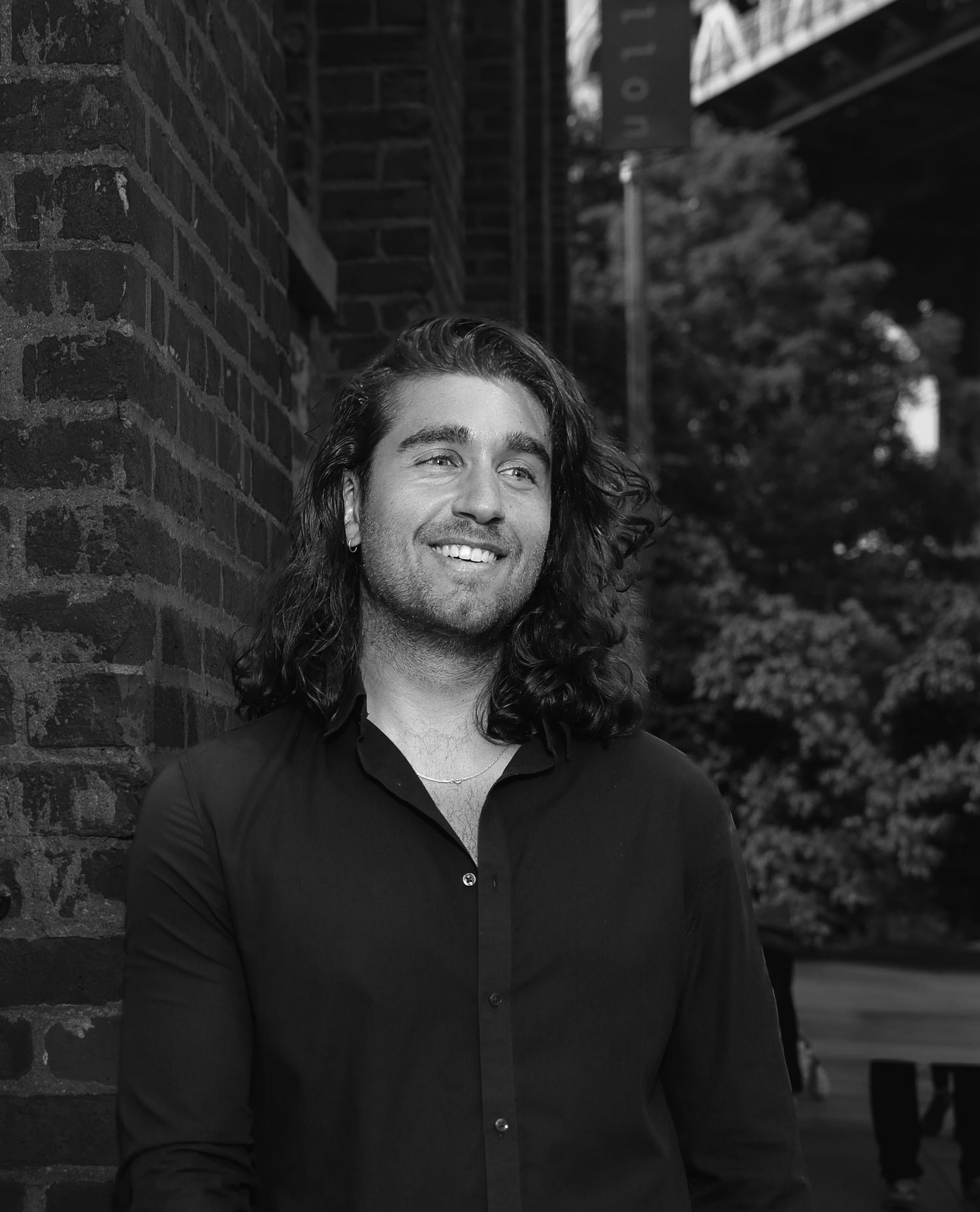 Smiling man with long curly hair in black shirt poses outdoors near brick wall in black and white photo.