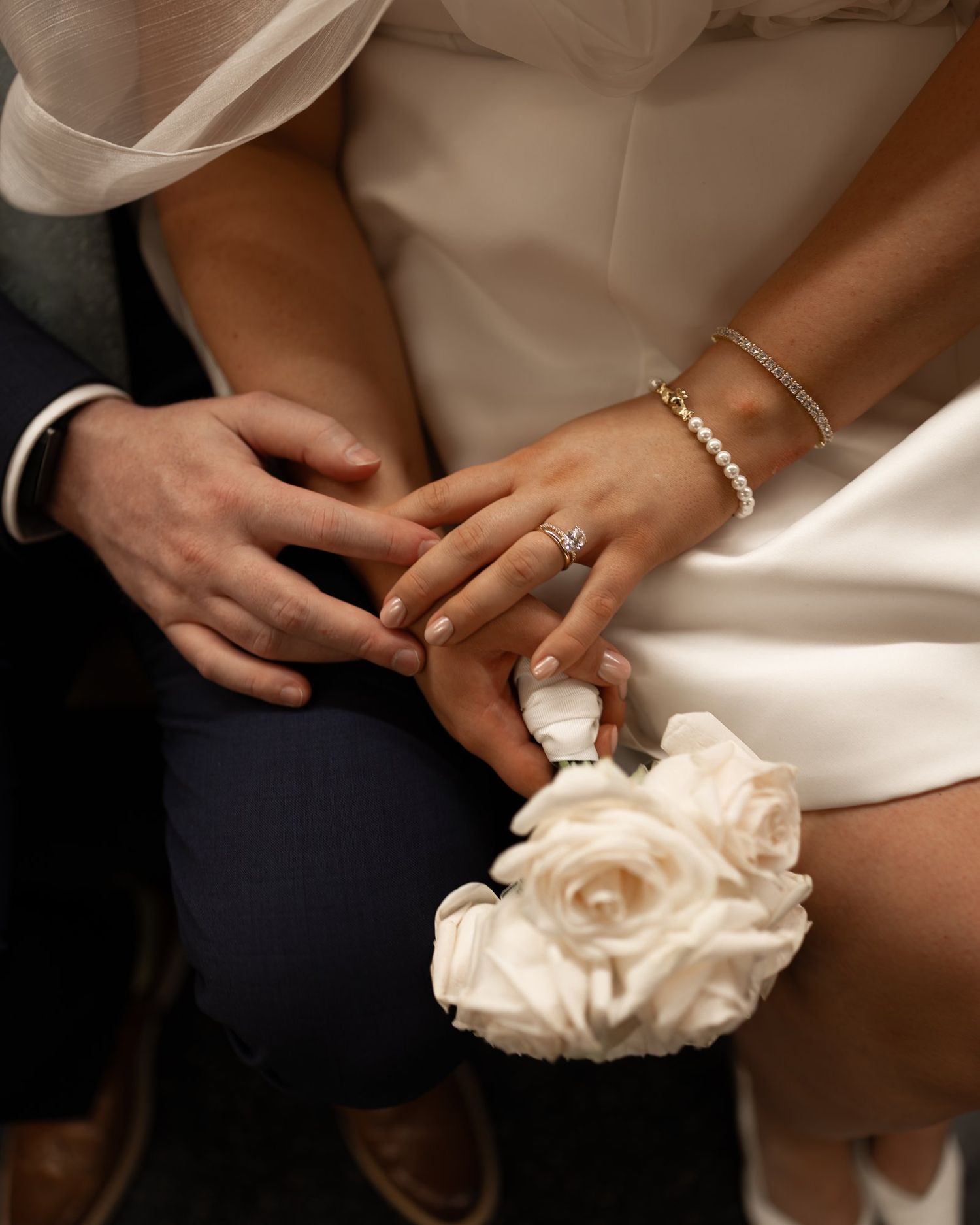 Close-up of bride and groom holding hands, showing wedding ring, white rose corsage, and gold bracelets.