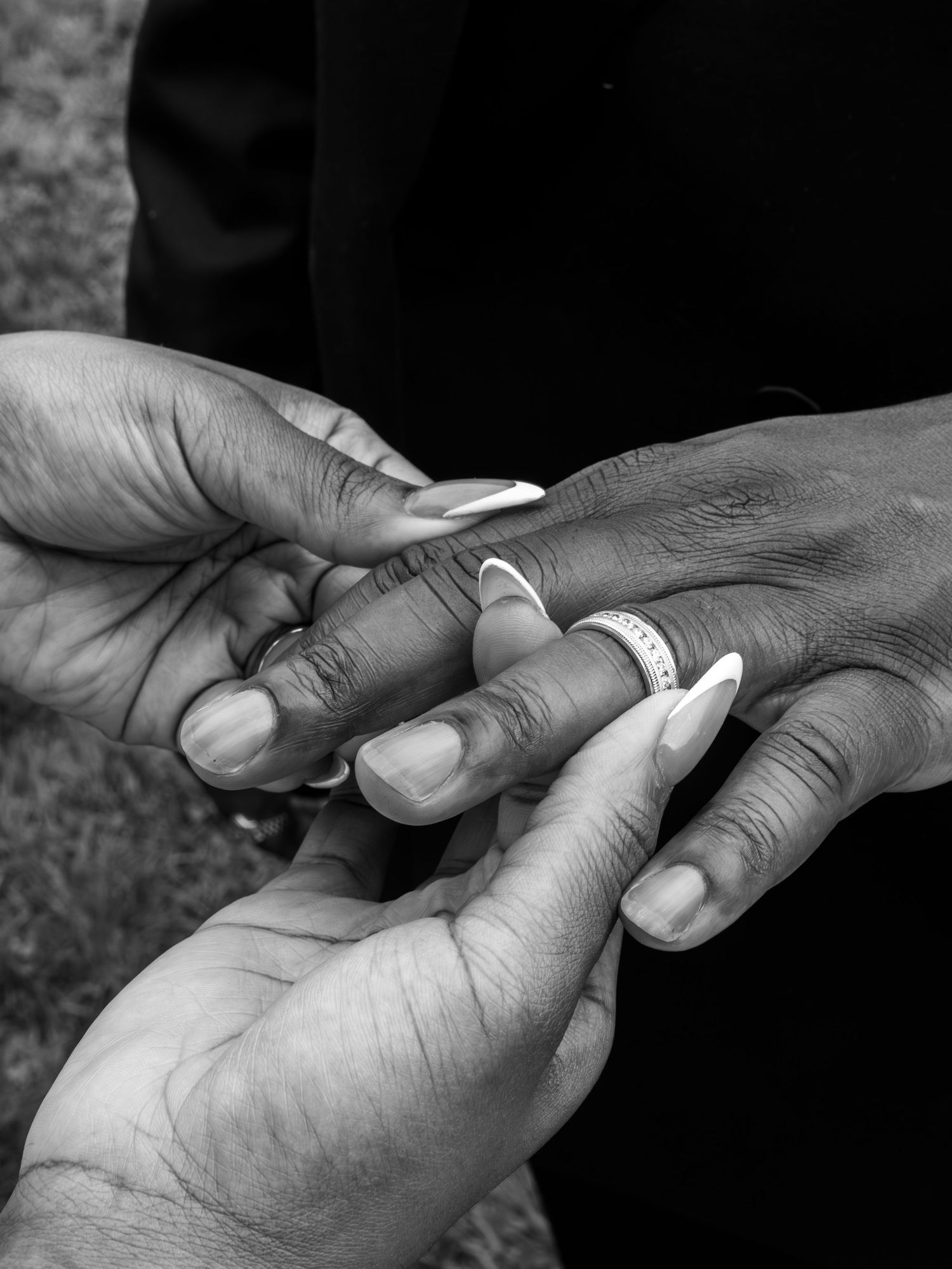 Black and white photo of two people exchanging a wedding ring during a ceremony, close-up of hands with manicured nails.