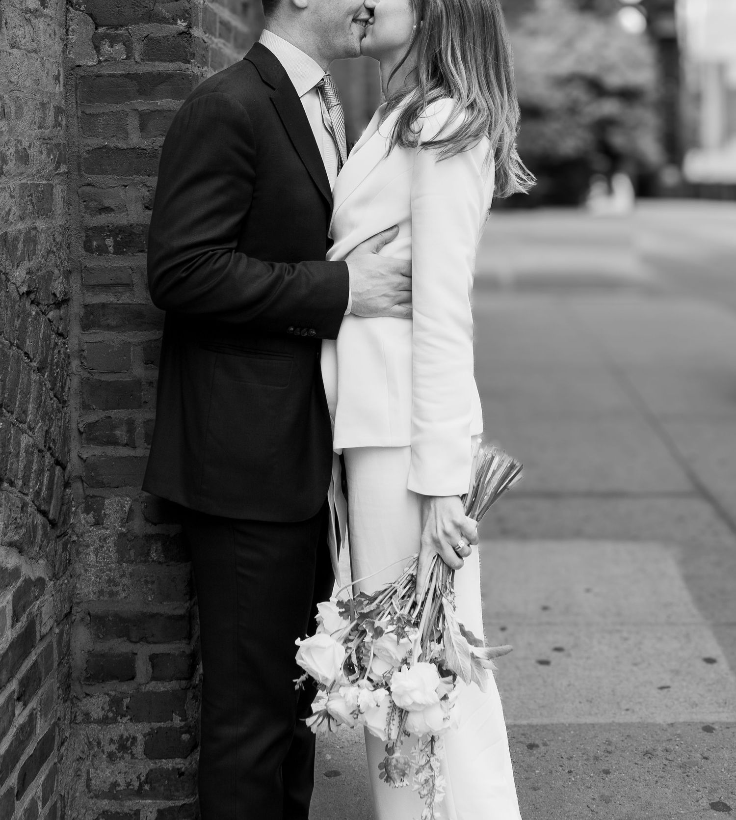Black and white photo of a couple sharing a kiss on a city sidewalk, bride holding a bouquet in a white suit.