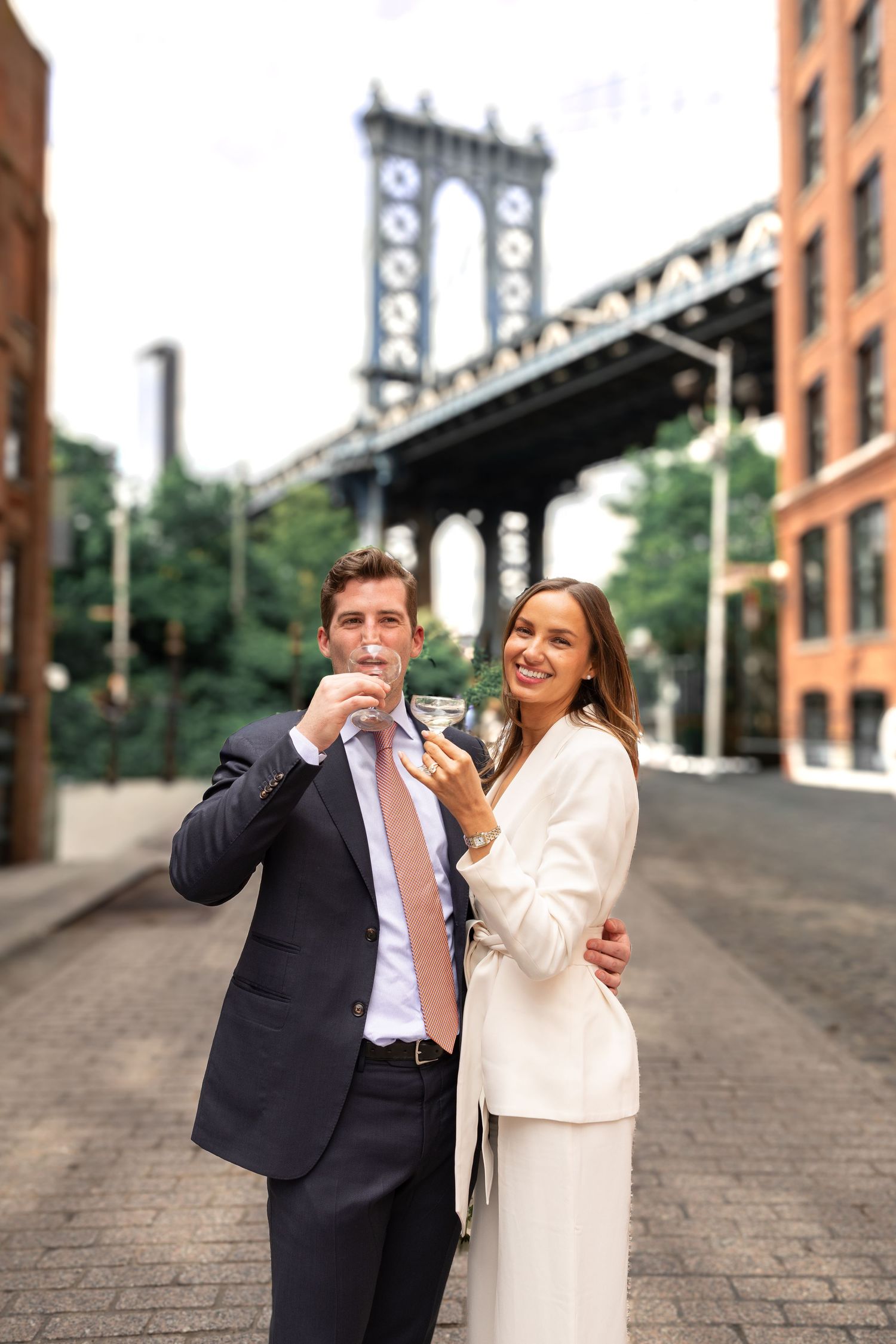Couple in formal attire toasting champagne on a cobblestone street with Manhattan Bridge in the background.