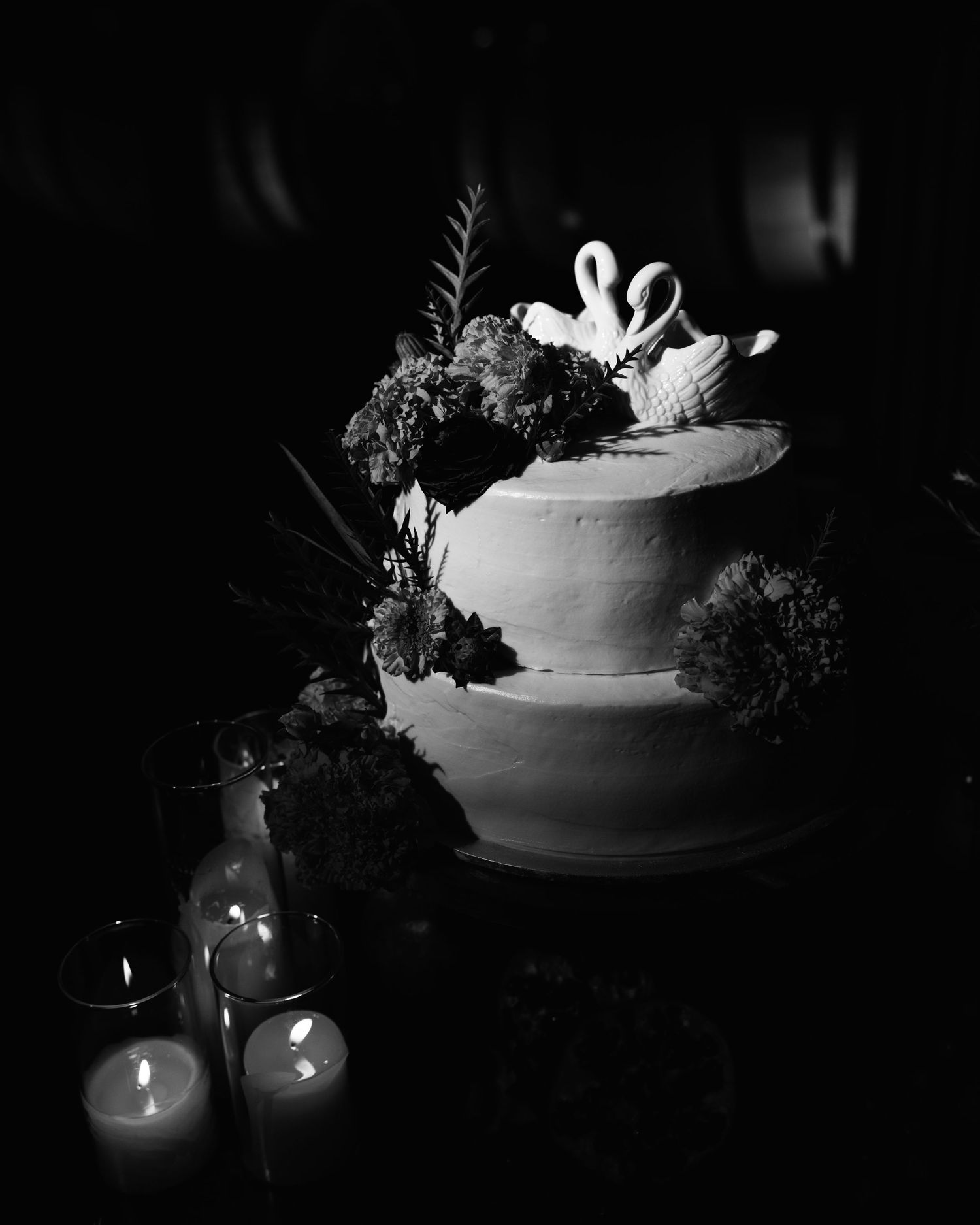 Black and white photo of a two-tier wedding cake topped with porcelain swans and flowers, glowing beside lit candles in a dark room.