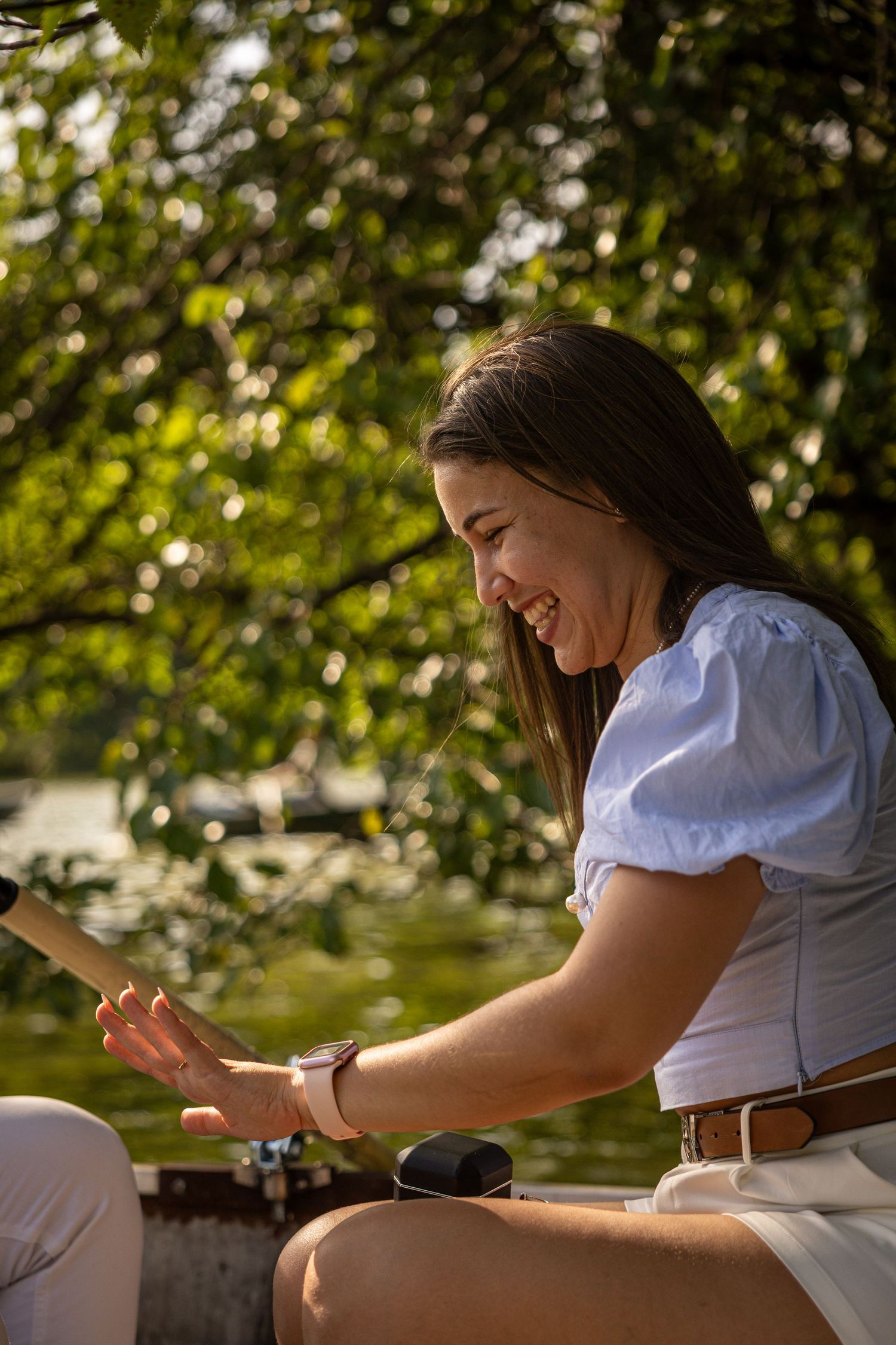 Smiling young woman in white blouse sitting outdoors near water, enjoying sunny day among green trees.