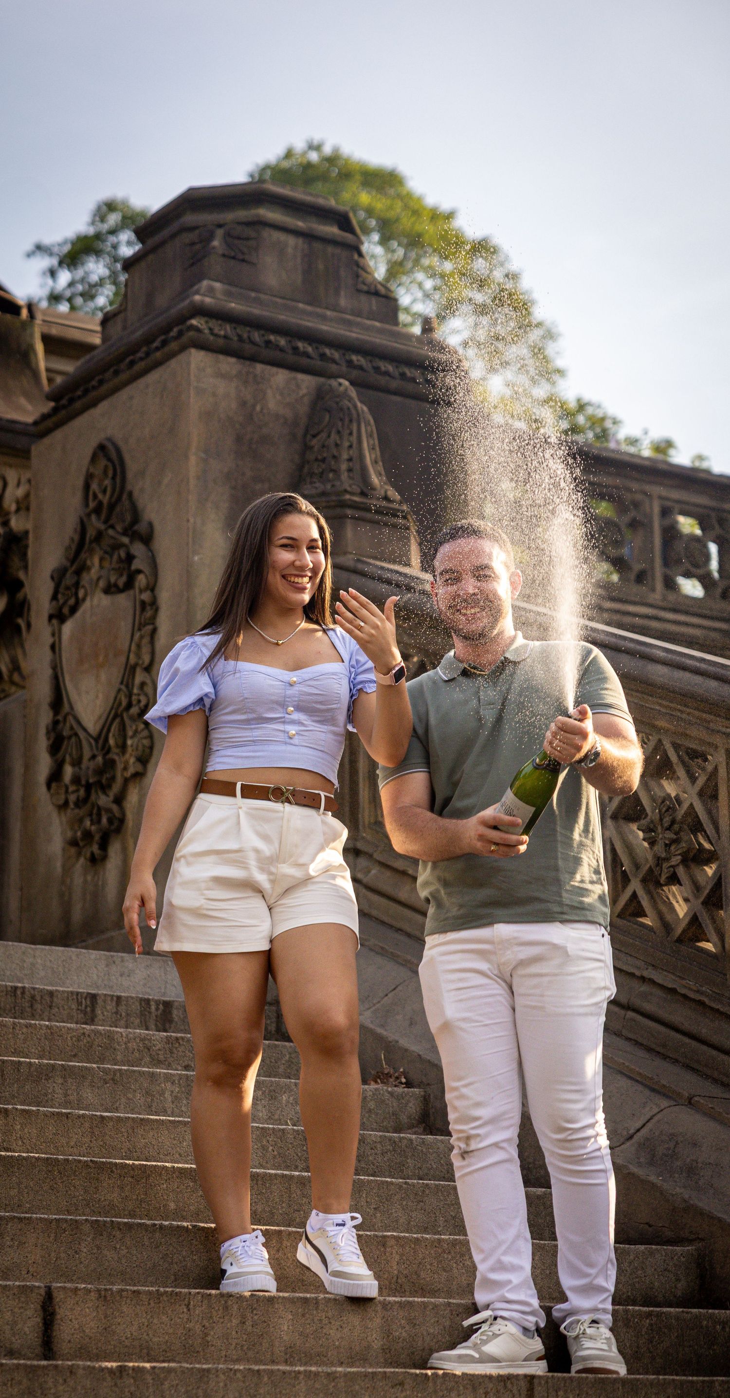 Couple celebrating an engagement on stone steps, woman showing ring while man pops champagne bottle outdoors.