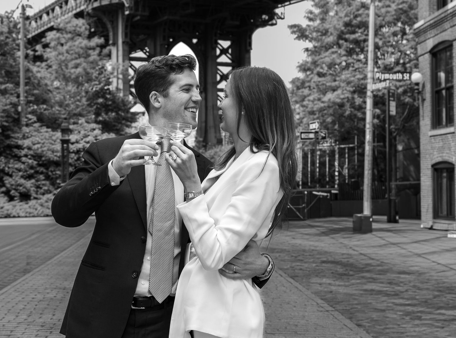 Black and white photo of a couple sharing a romantic moment on a city street near a bridge.
