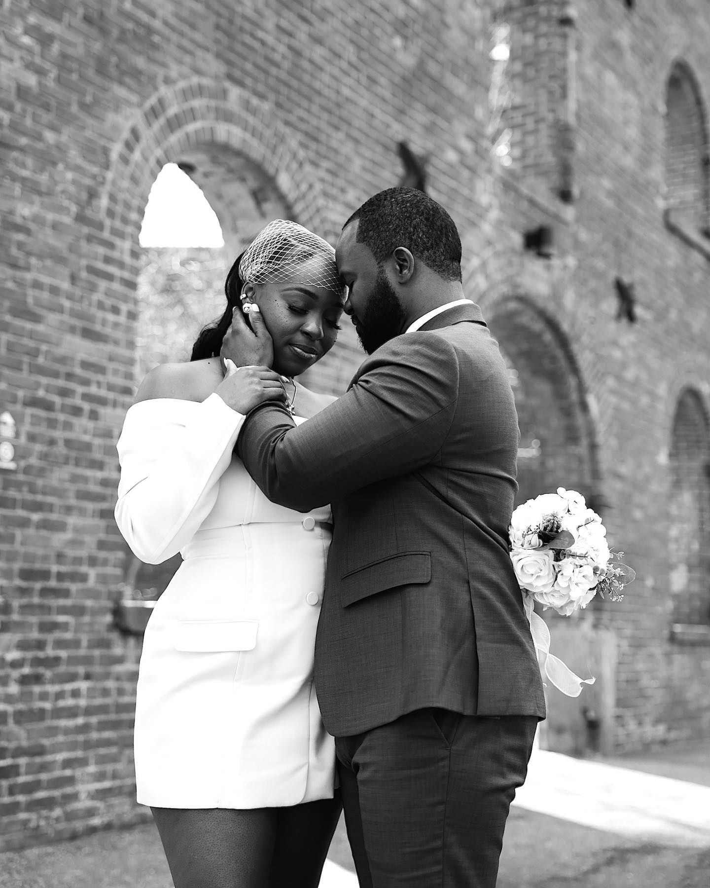 Black and white photo of a couple in wedding attire sharing an intimate moment against a brick wall backdrop.