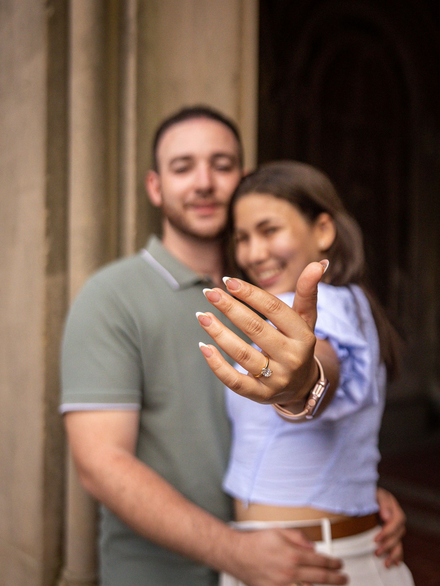 Happy couple showing off engagement ring, woman extending hand toward camera with diamond ring, man embracing her from behind.