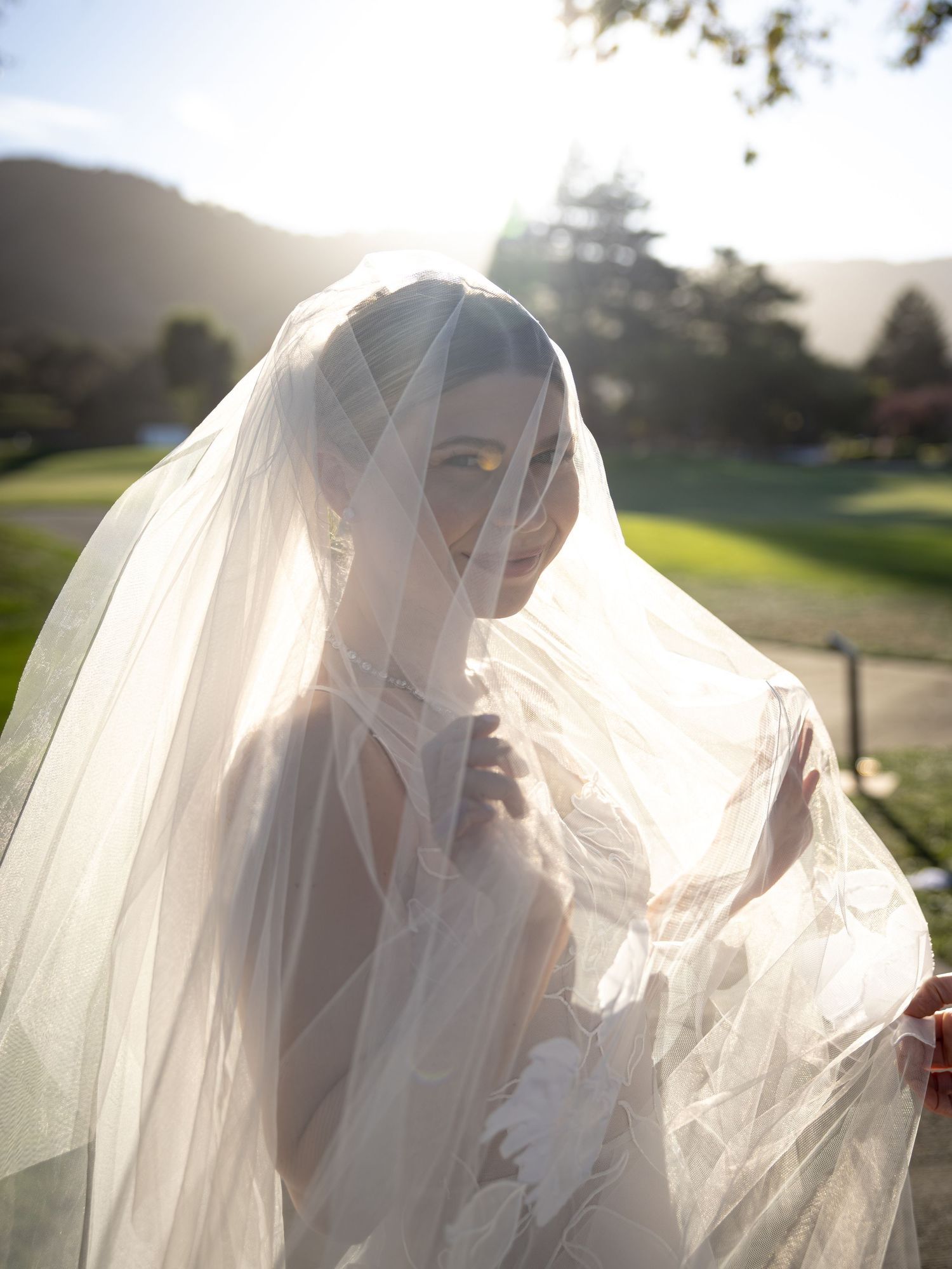 Radiant bride smiling softly beneath her flowing sheer veil in golden sunlight at an outdoor wedding in California.
