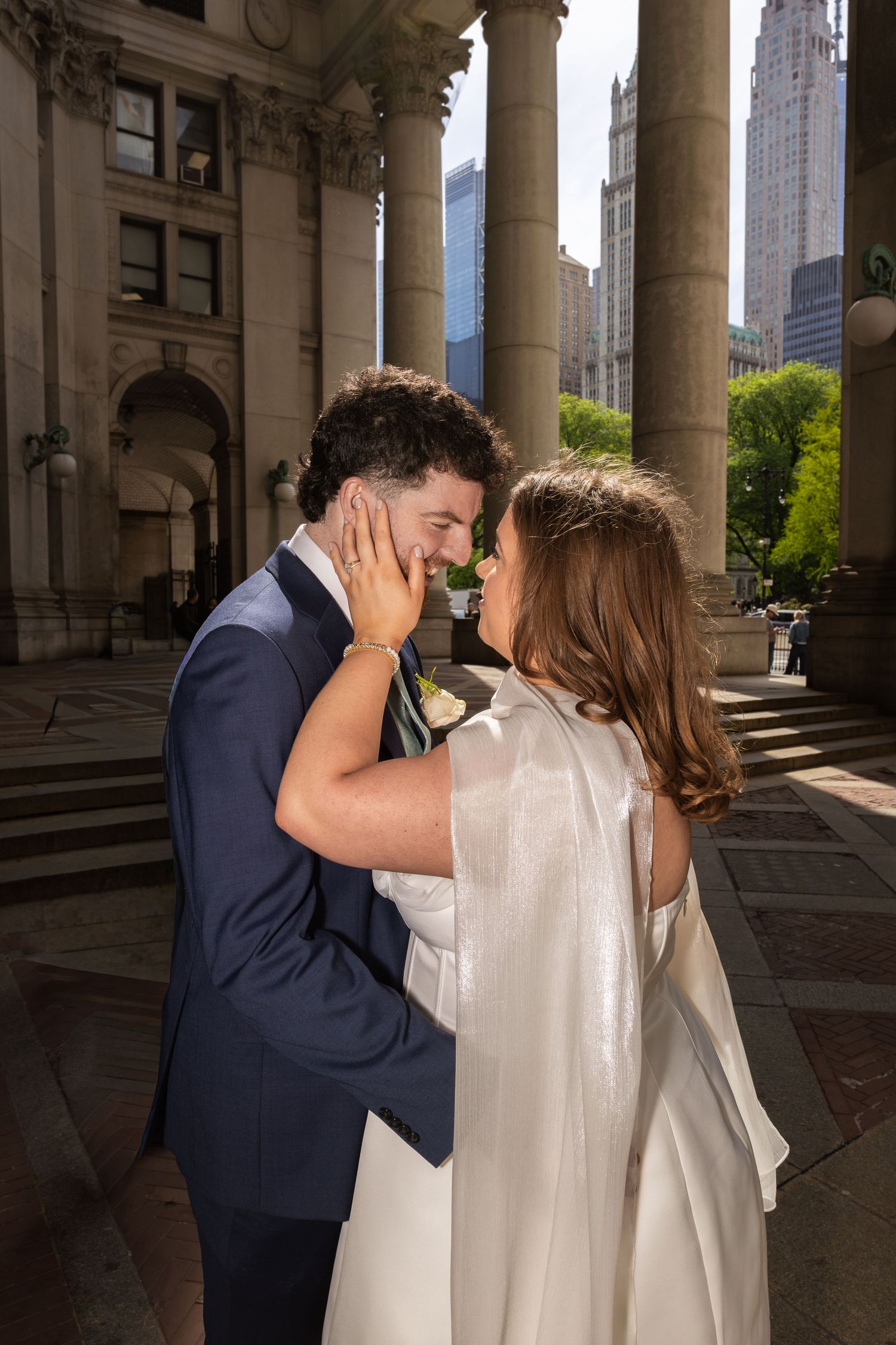 Bride and groom share a romantic moment outside a grand NYC building, she in a white cape dress, he in a navy suit.