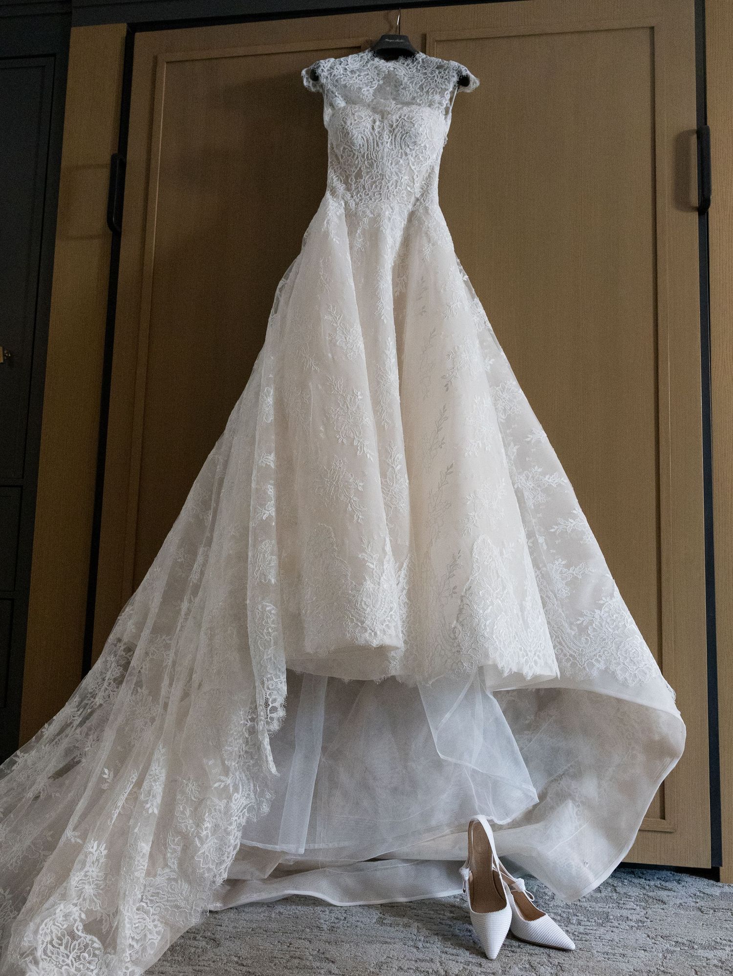 Elegant lace wedding gown with a long train hanging on a door above white bridal heels in New Jersey.