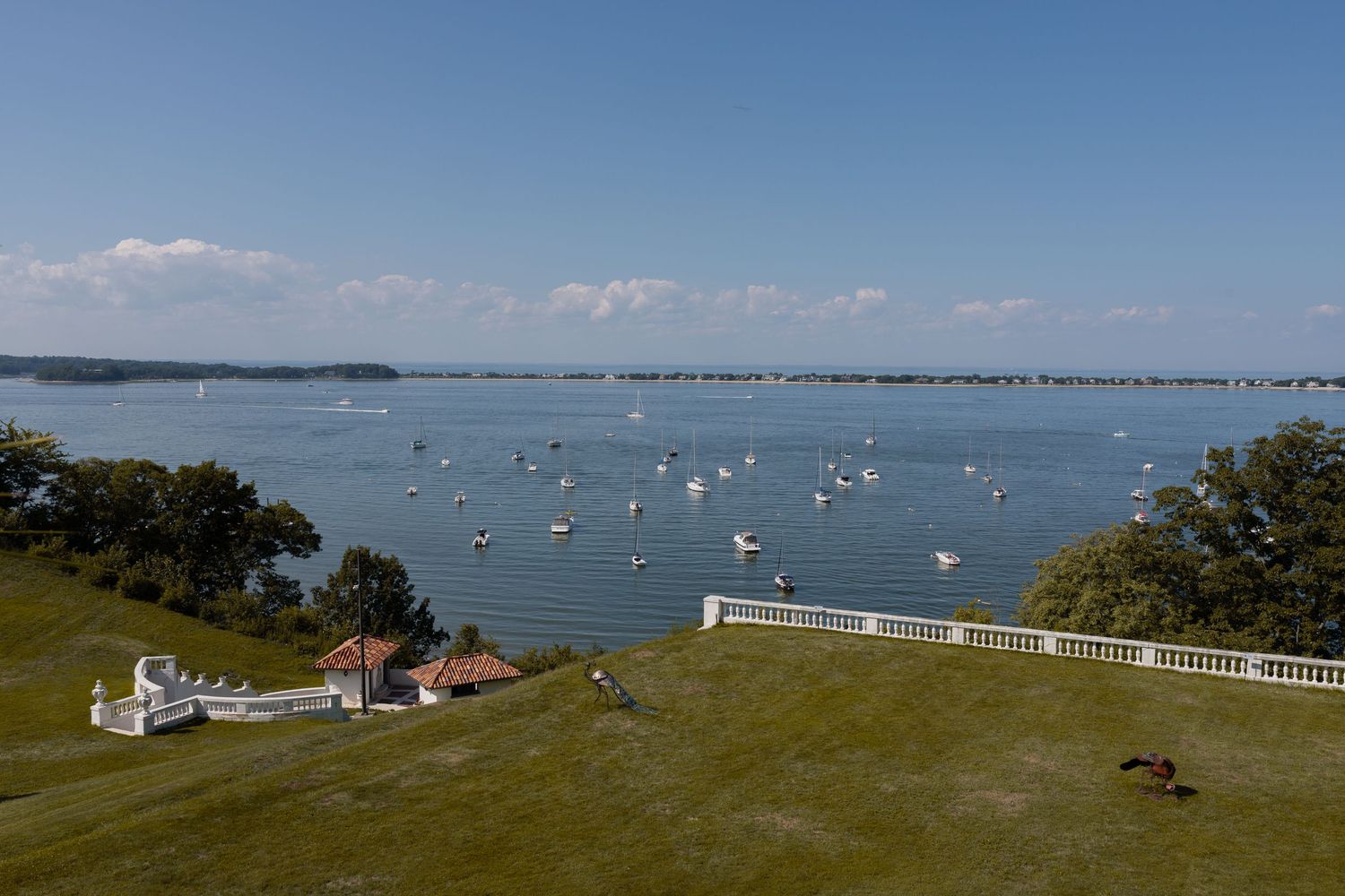 Aerial view of a vast bay with sailboats, lush green lawn, and a white fence on a sunny day.