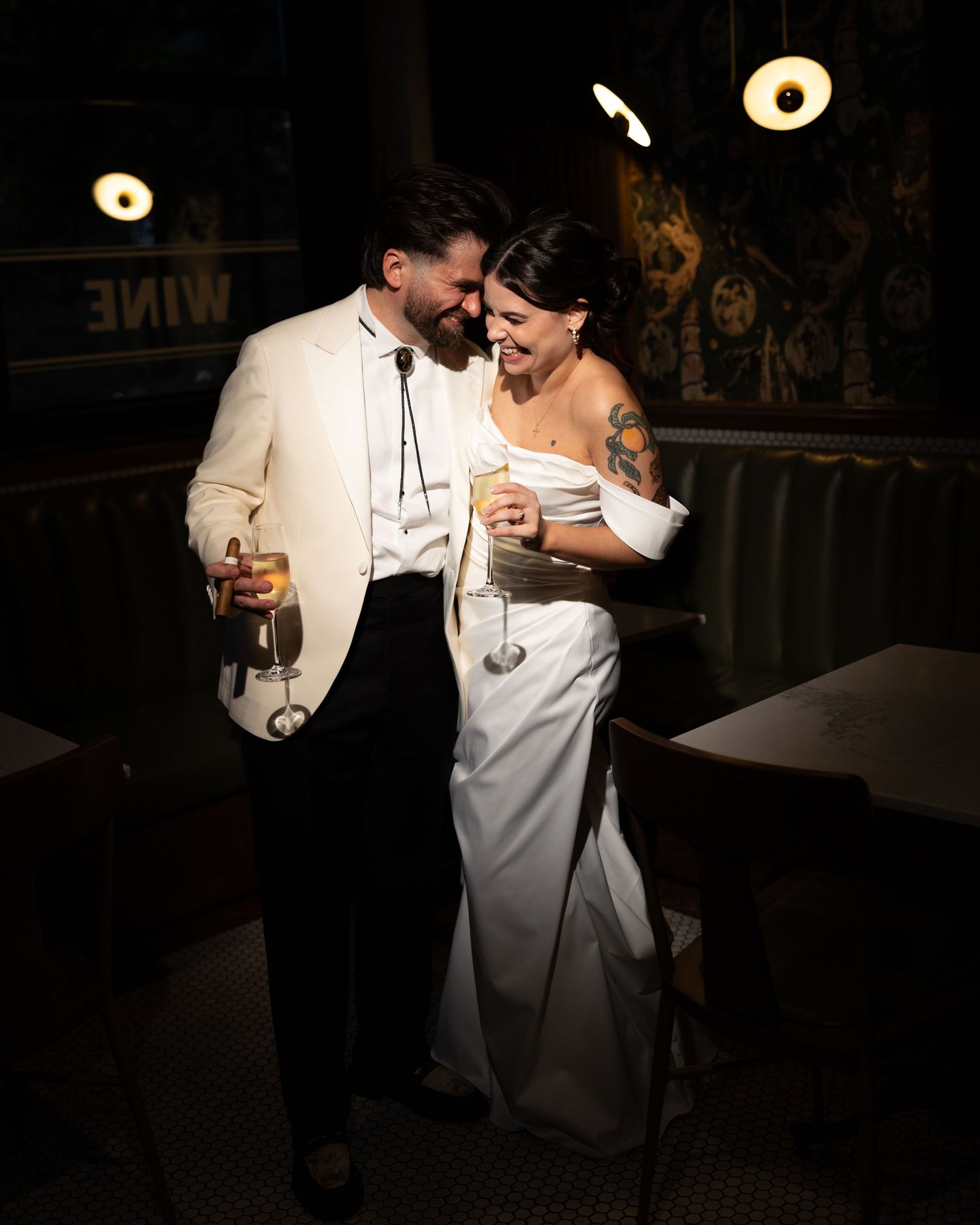 Smiling bride in a white satin gown and groom in a cream jacket hold champagne glasses in a moody, dimly lit wine bar.