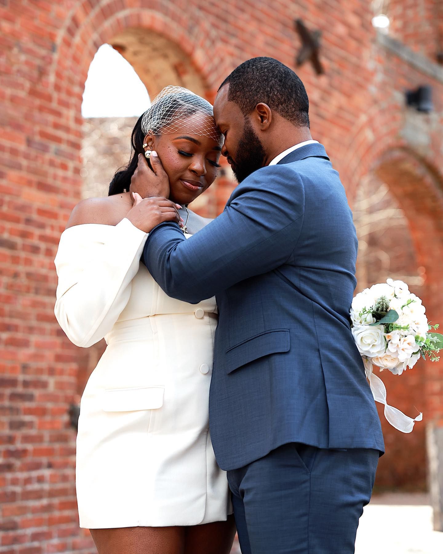 A couple shares an intimate moment in wedding attire, groom in blue suit holding white flowers, bride in white mini dress, brick backdrop.