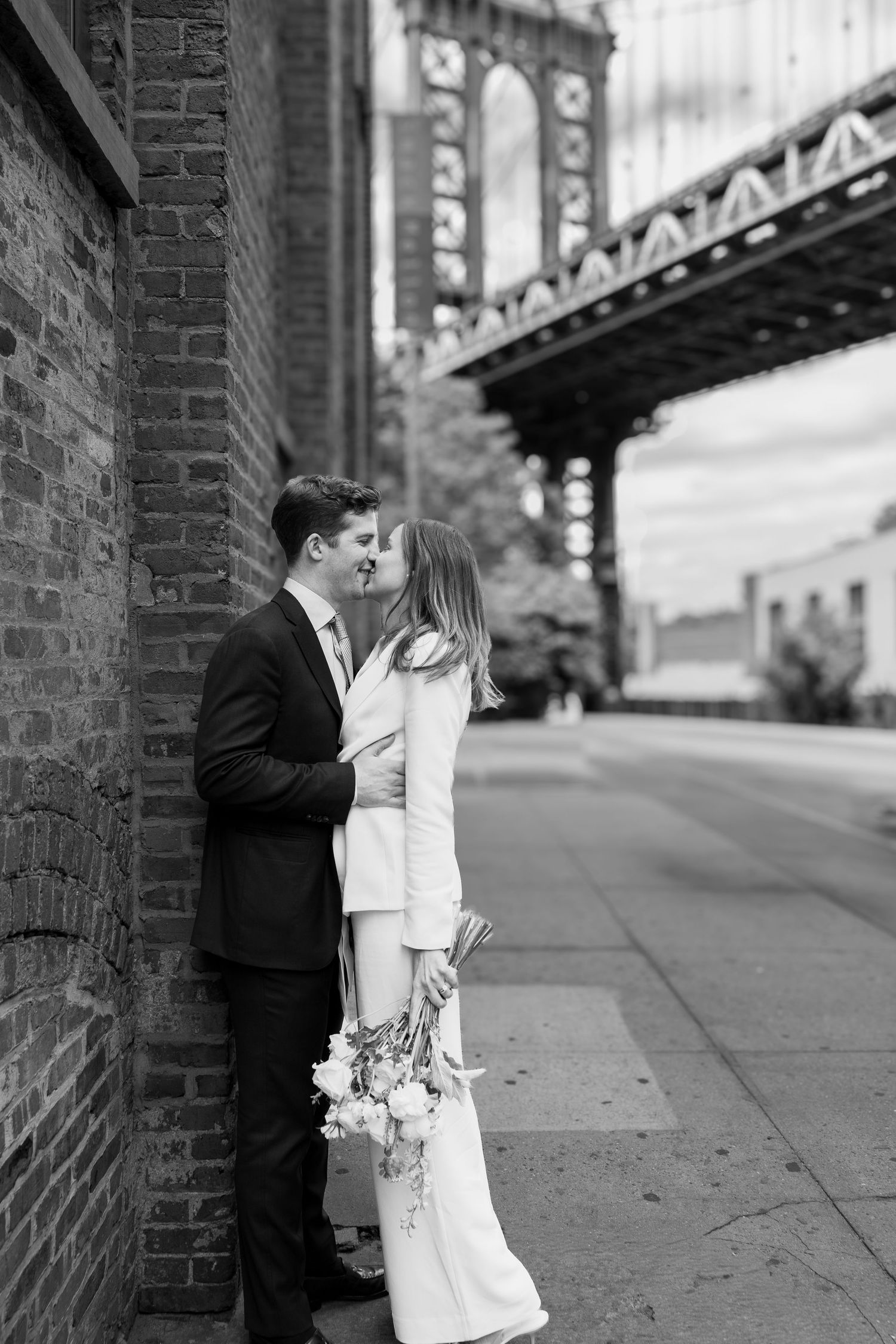 Couple kissing in wedding attire by brick wall with Manhattan Bridge in black and white NYC photo.