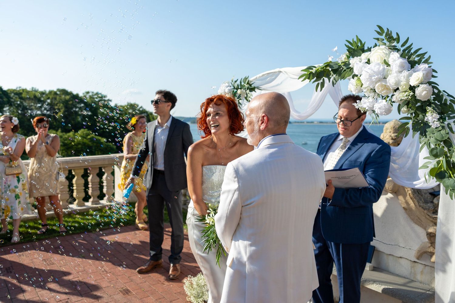 Outdoor seaside wedding ceremony with a bride and groom facing an officiant under a floral arch while guests watch from a terrace.