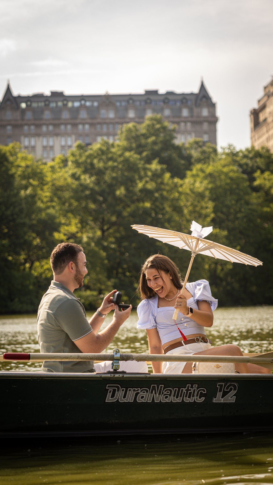 Couple on a DuraNautic rowboat in Central Park, woman holding parasol while man photographs her, NYC buildings behind.