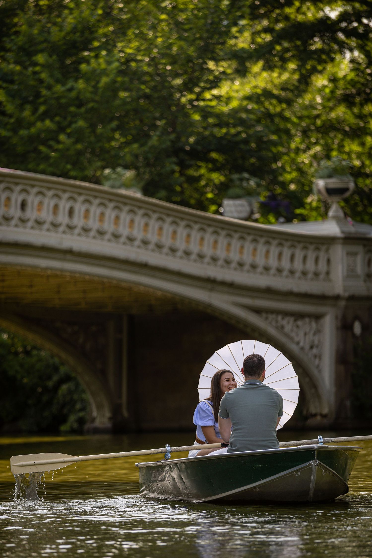 Couple rowing a boat under Bow Bridge in Central Park, sharing a white parasol umbrella.