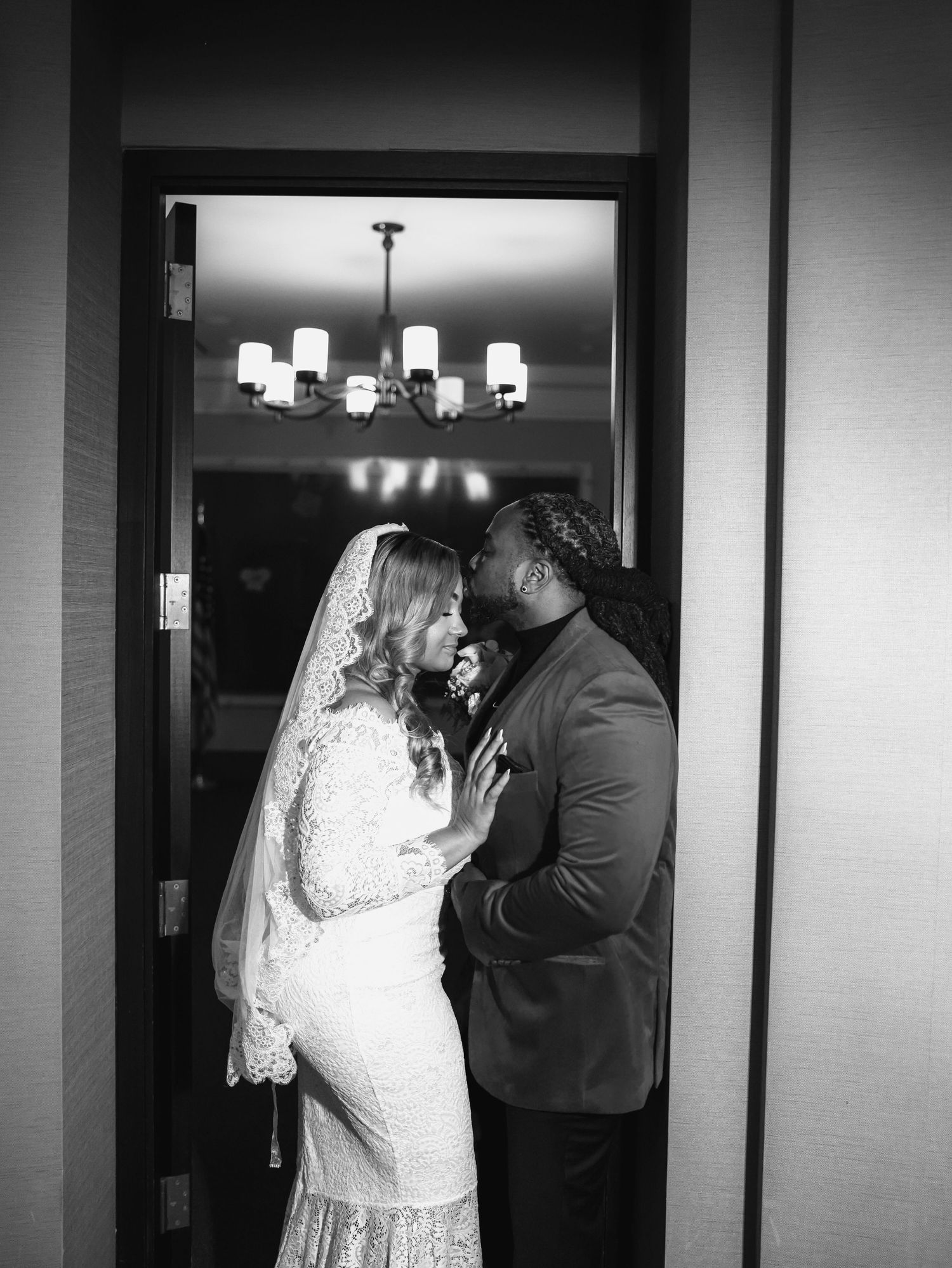 Black and white wedding photo of bride in lace gown and groom in suit sharing intimate moment in doorway.