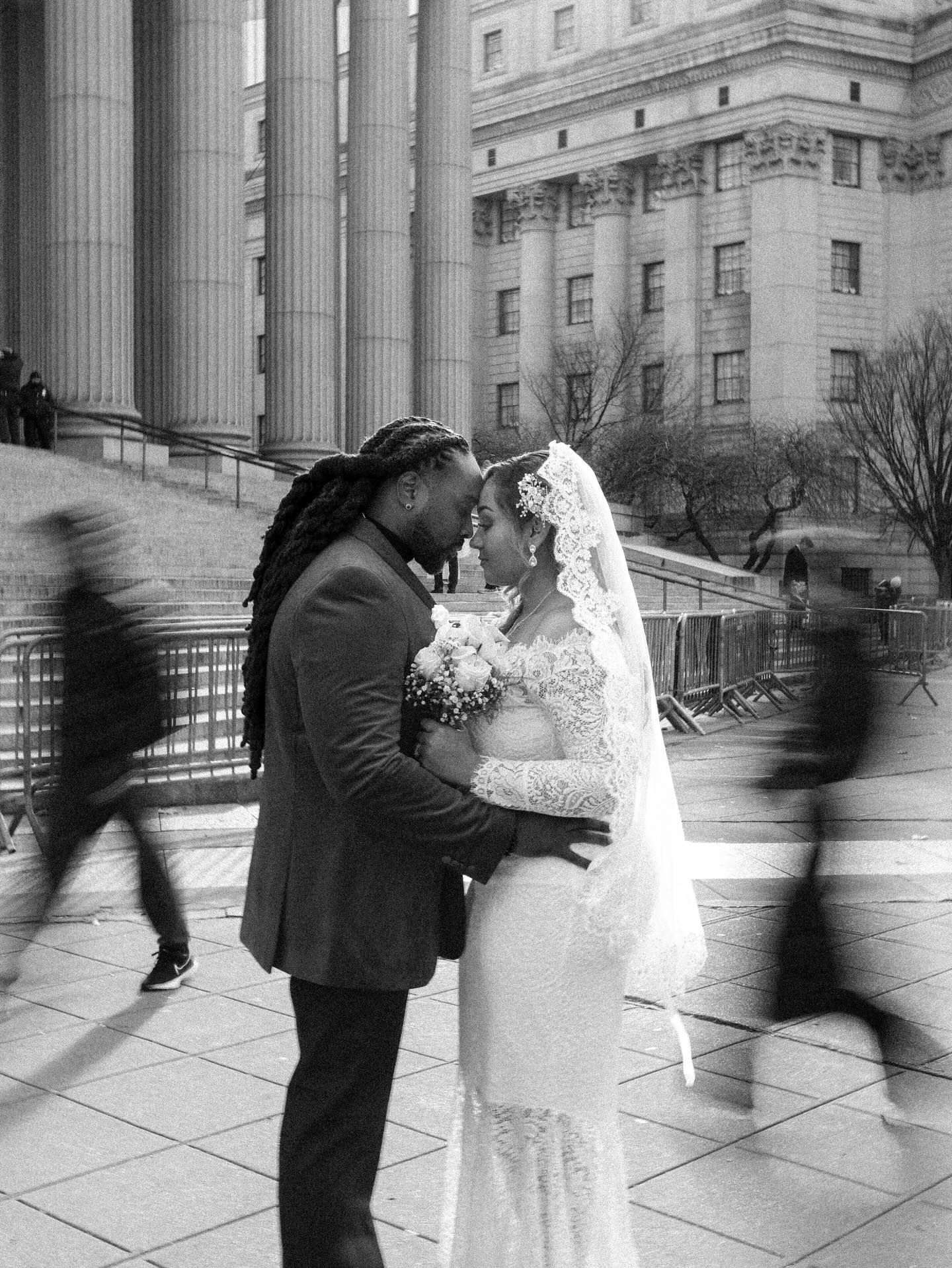Black and white photo of a couple in wedding attire embracing near grand columns with blurred pedestrians passing by.