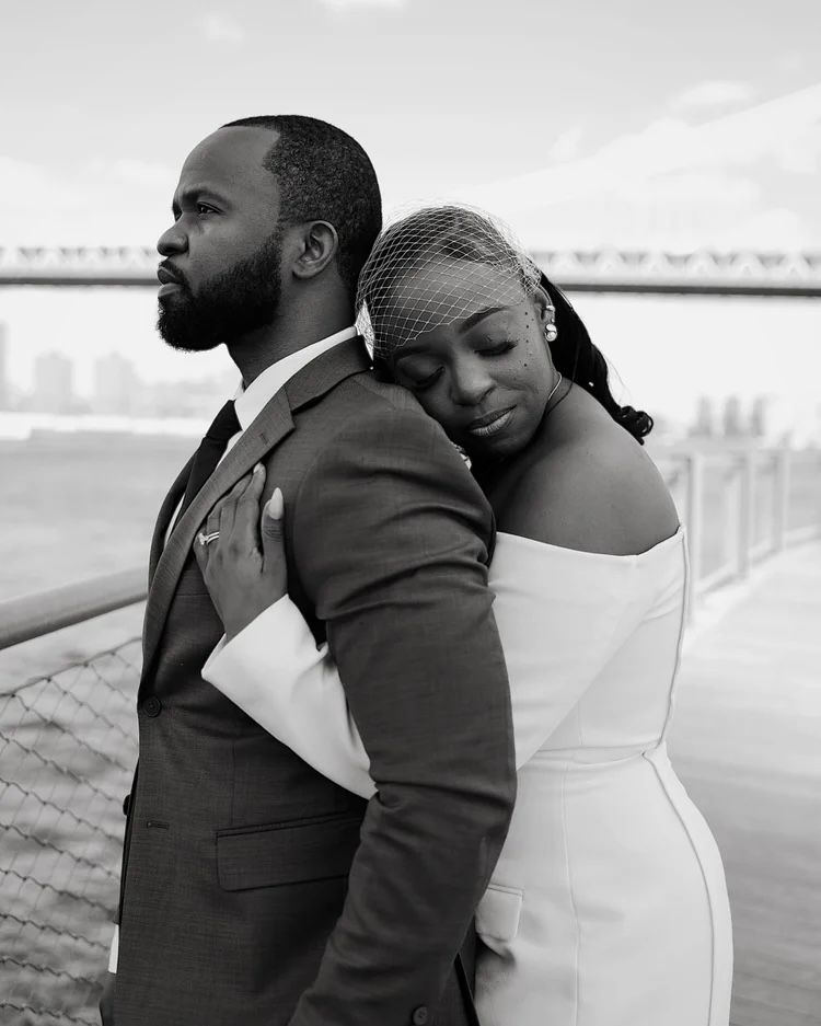 Black and white photo of a bride leaning on groom's back by waterfront bridge in Brooklyn, New York City.