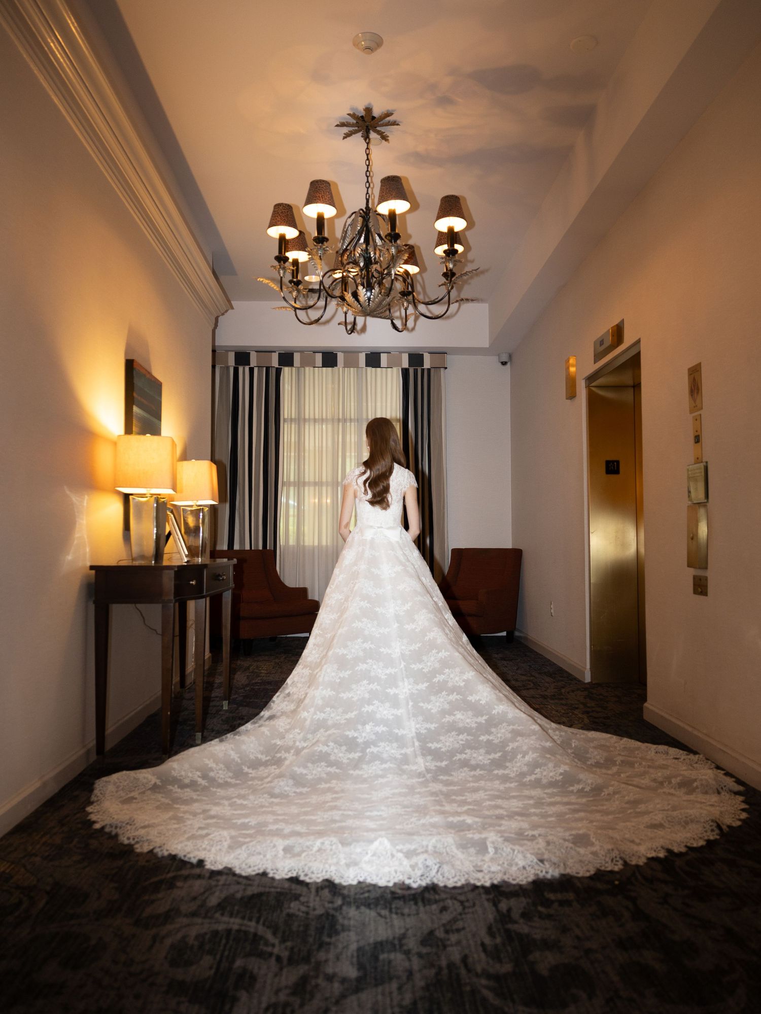 Bride in cathedral lace gown standing in chandelier-lit hotel hallway from behind In New Jersey.