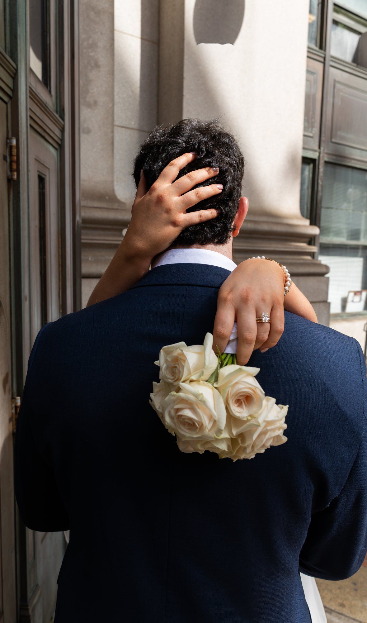 Bride holding white roses bouquet embracing groom in navy suit from behind at elegant building exterior.