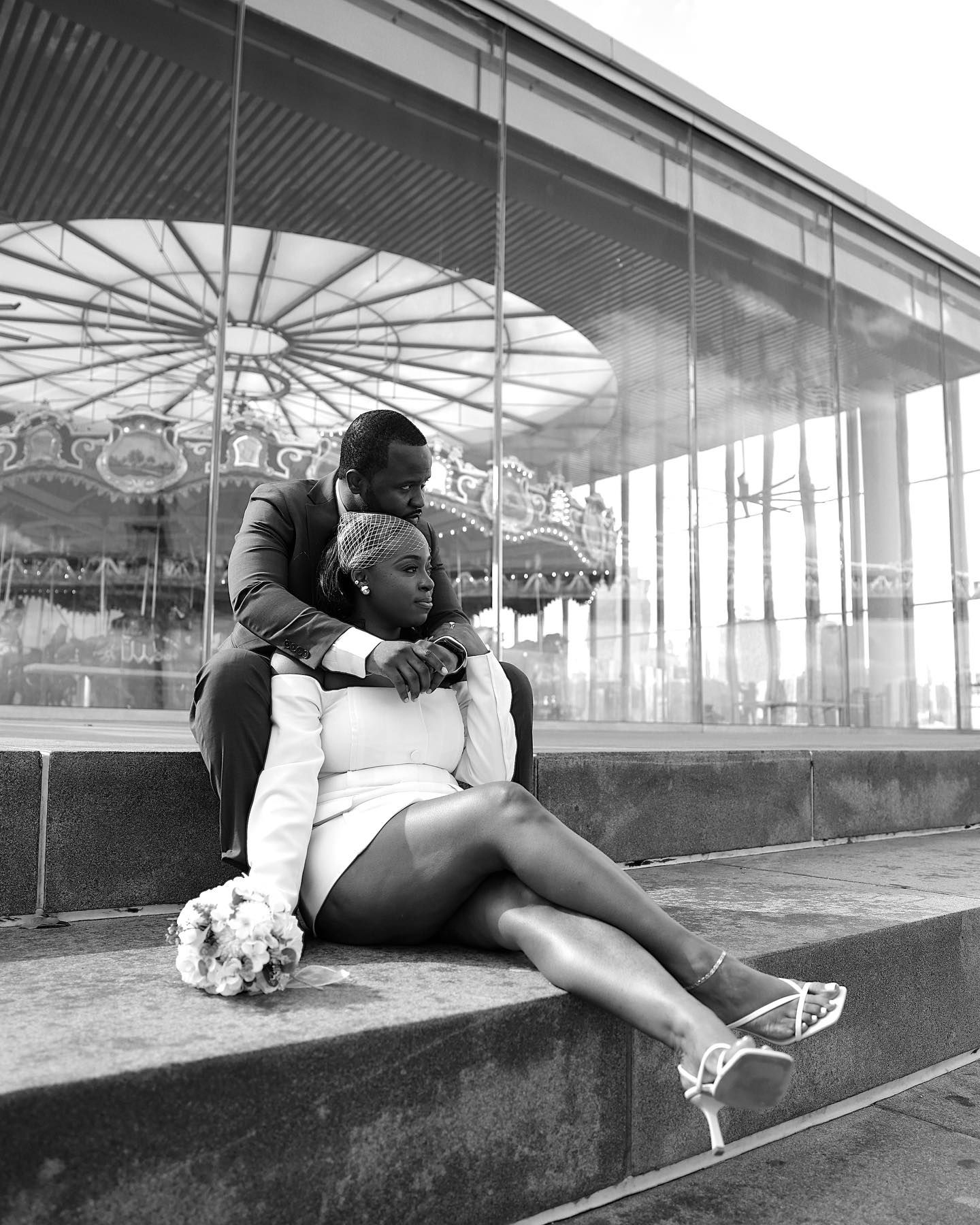 Black and white photo of a couple in wedding attire sitting on steps near a carousel, sharing a tender romantic moment.