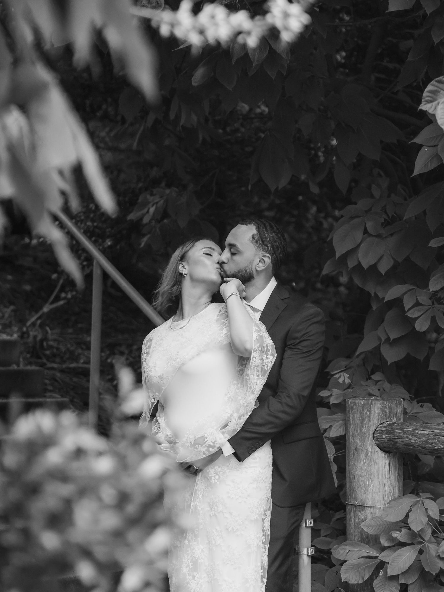 Black and white photo of a bride and groom sharing a romantic kiss surrounded by lush green foliage.